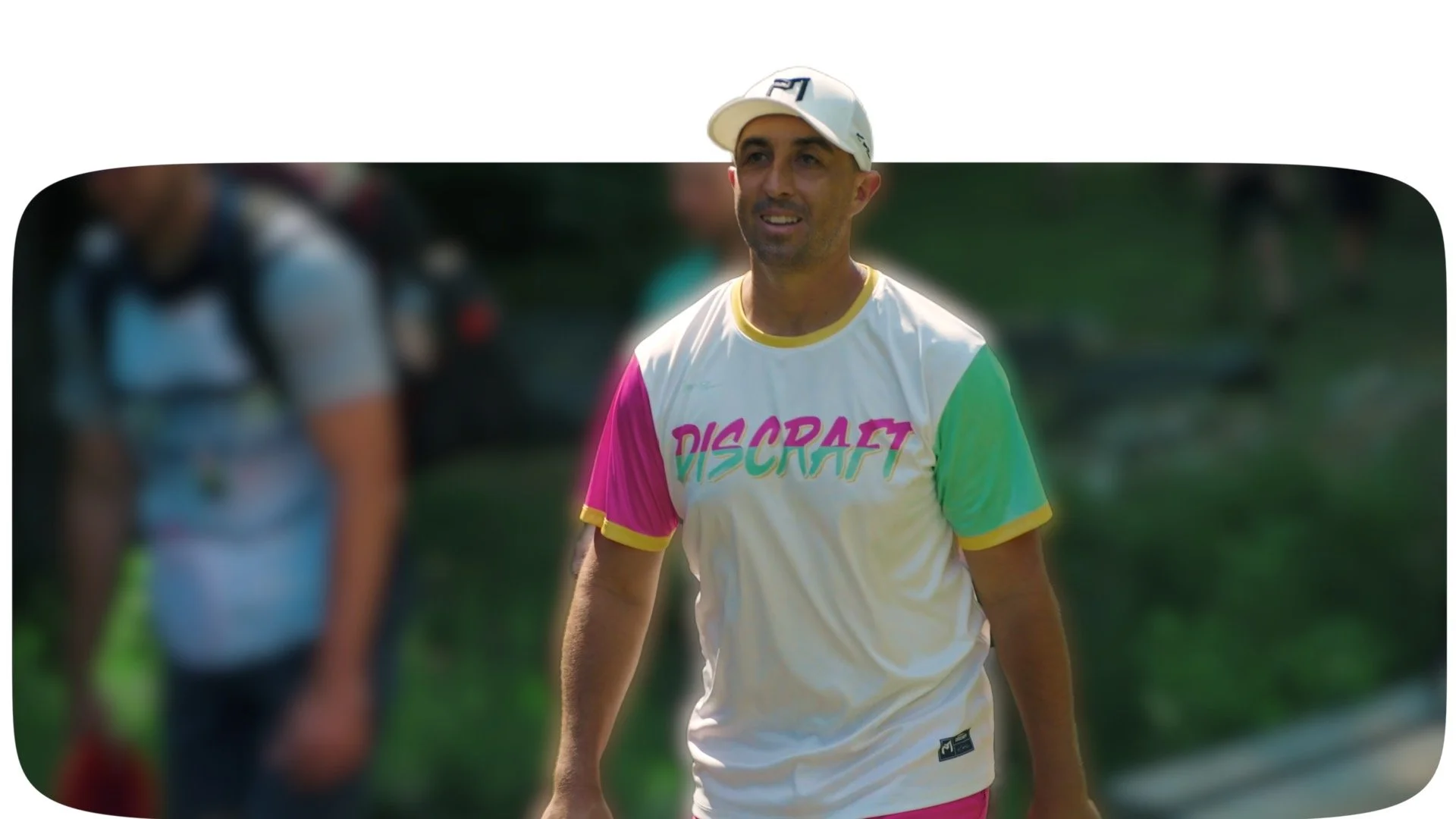 A man wearing a cream-colored cap and a colorful t-shirt with the logo 'DISCRAFT' is standing outdoors on a sunny day, with greenery in the background.