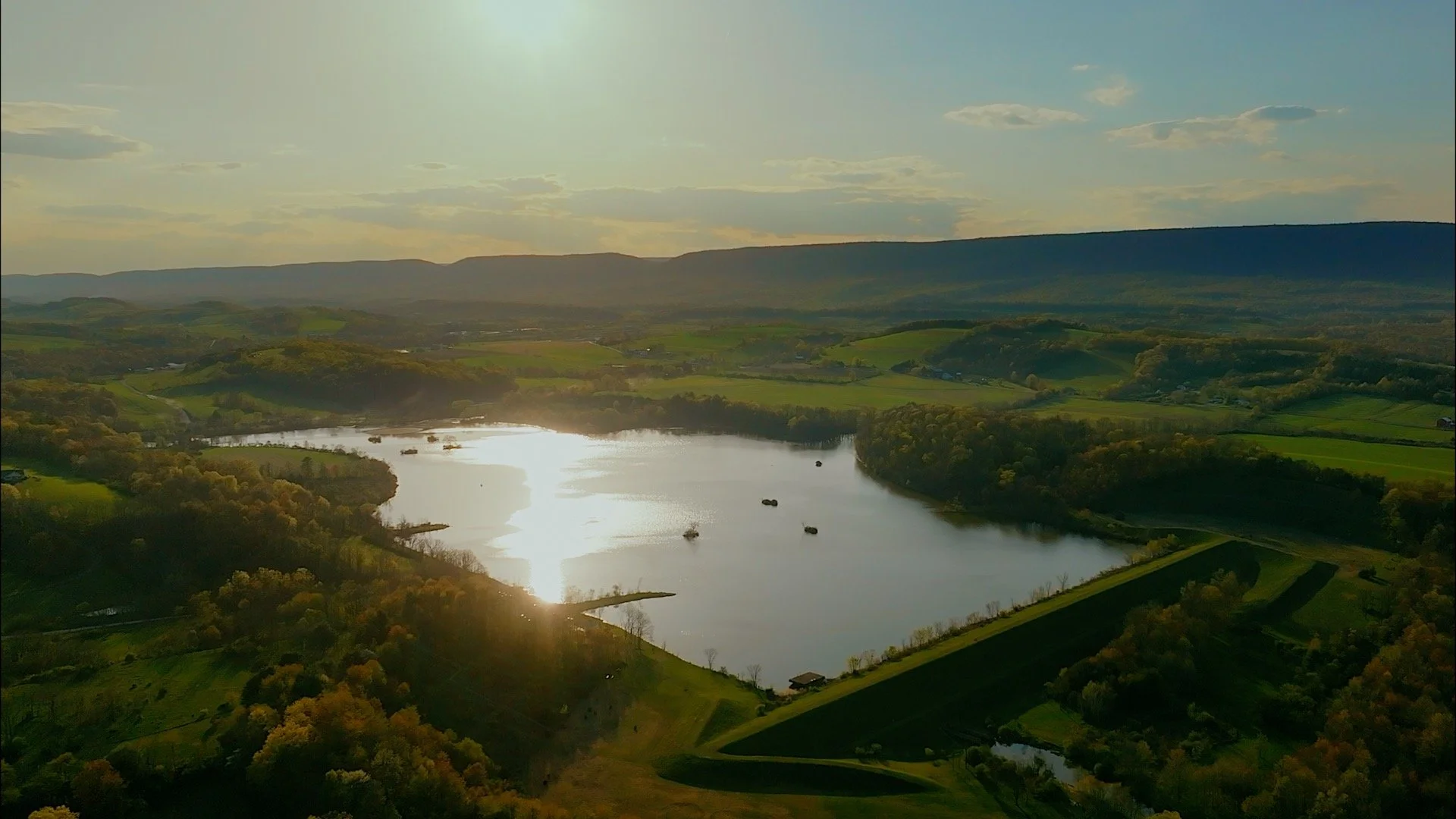 An aerial view of a disc golf courses lake surrounded by green hills and fields, with boats on the water and a bright sun reflecting off the lake surface.