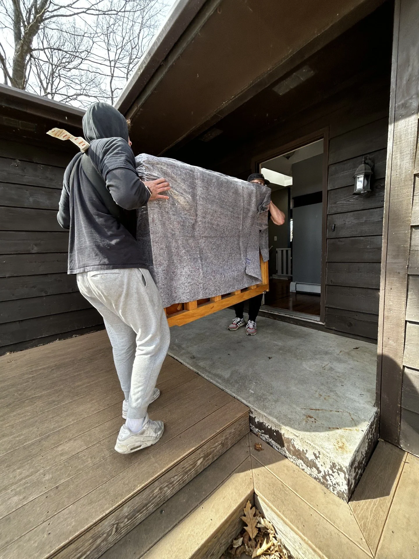 Two men are carrying a large piece of furniture wrapped in grey fabric into a house.