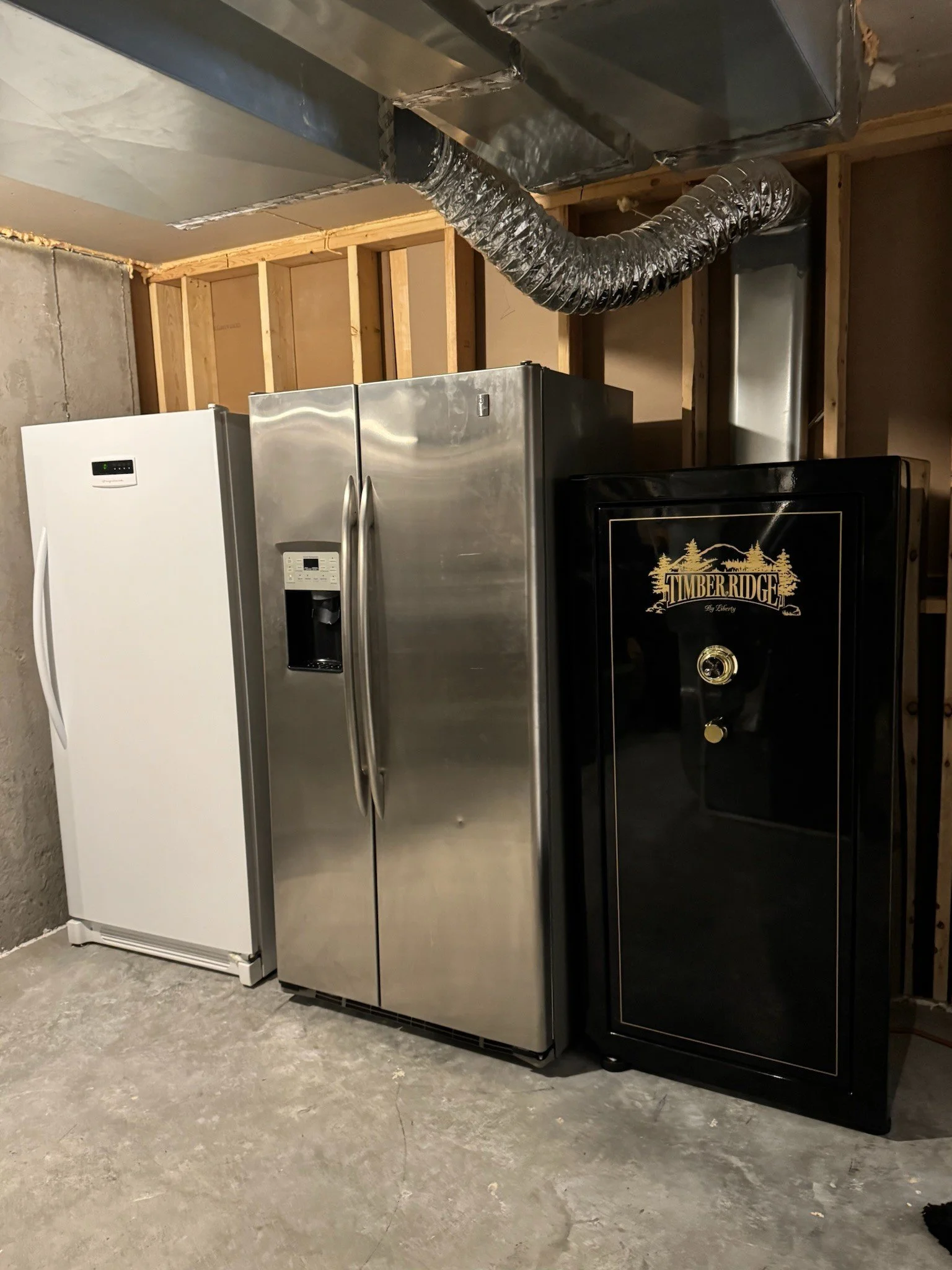A white refrigerator, a stainless steel refrigerator, and a black safe are placed on a concrete floor in an unfinished basement with wooden framing and ductwork overhead.
