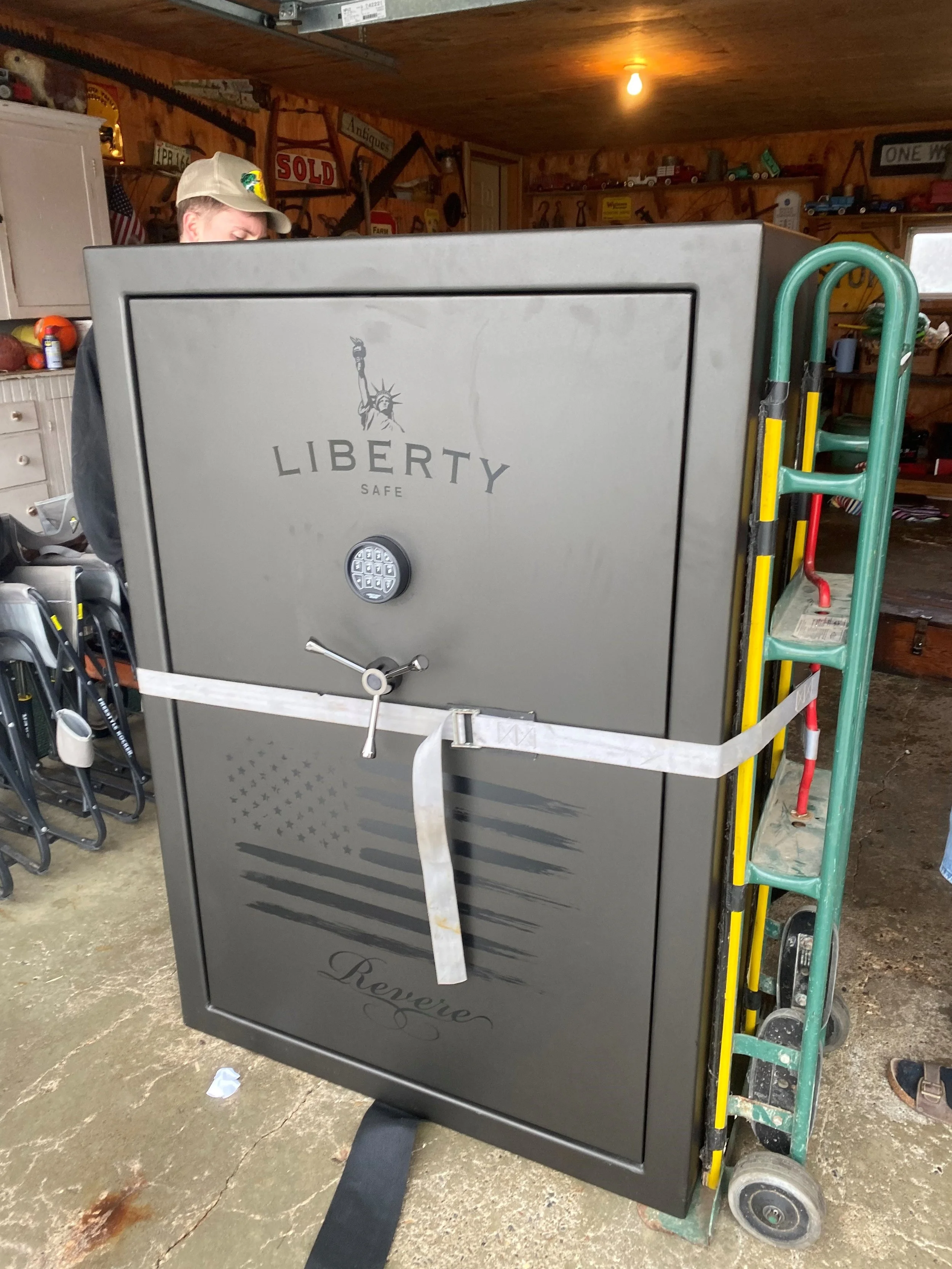 A large, grey Liberty Safe with a keypad lock and an American flag graphic, secured with a white strap on a green hand truck in a garage.