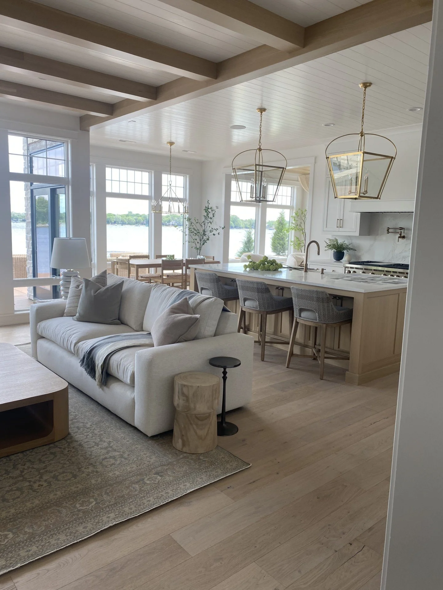 Bright open-concept living space with a white sofa, a beige accent stool, a wooden coffee table, and a view of the kitchen and dining area with large windows overlooking a lake.