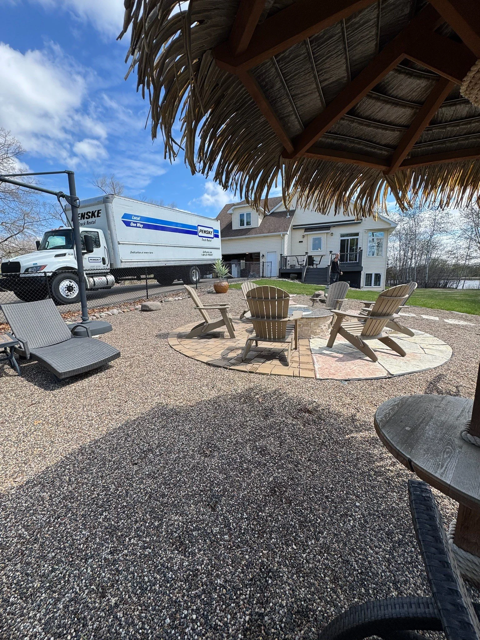 Outdoor backyard scene with Adirondack chairs around a fire pit, gravel ground, a thatched gazebo overhead, a large house in the background, and a Penske moving truck parked near a fence.
