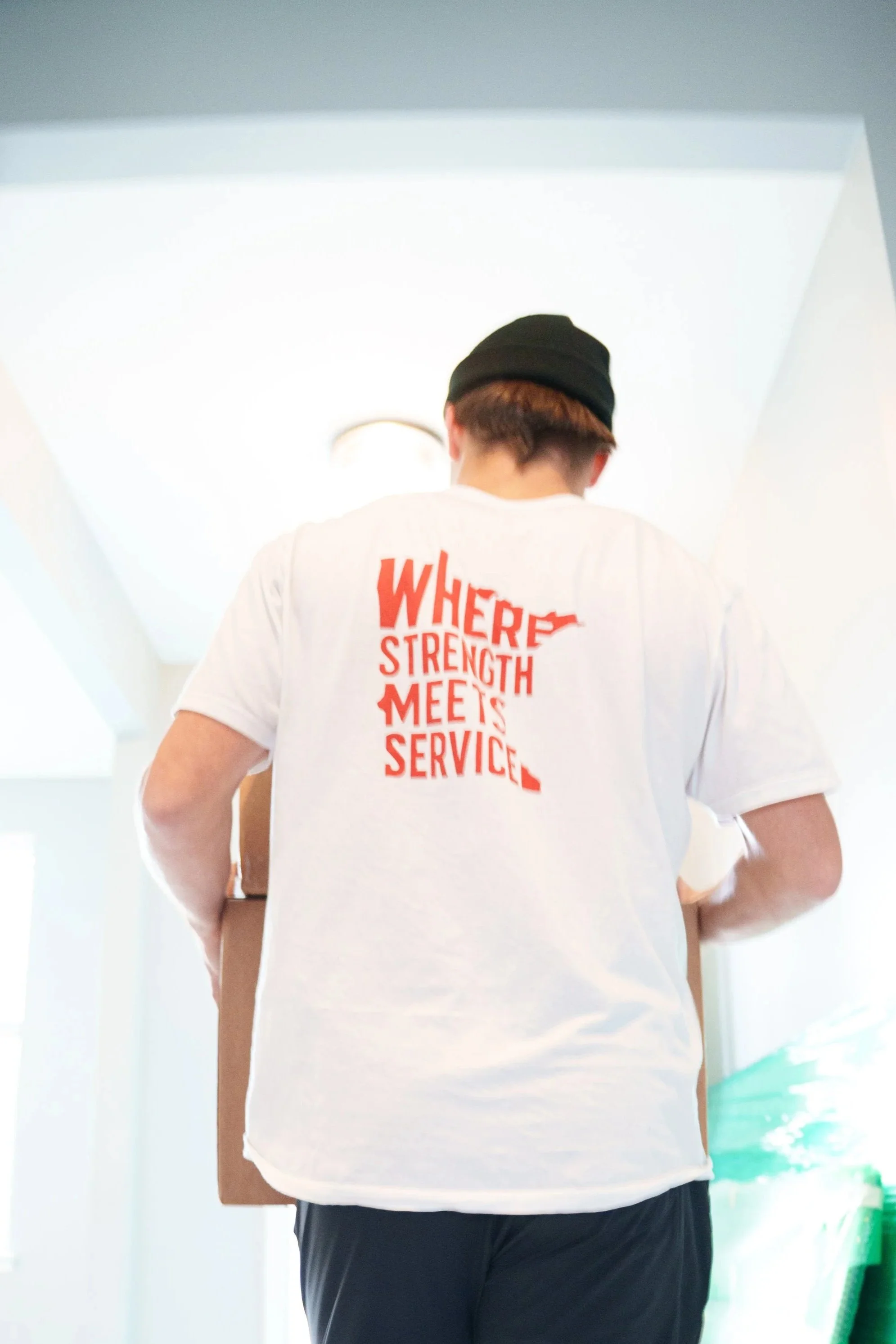 Two young men are unloading a mattress from a moving truck using a ramp on a suburban street. One is inside the truck helping to lift the mattress, while the other is outside on the ramp, assisting with the move. The background shows trees, parked cars, and houses.