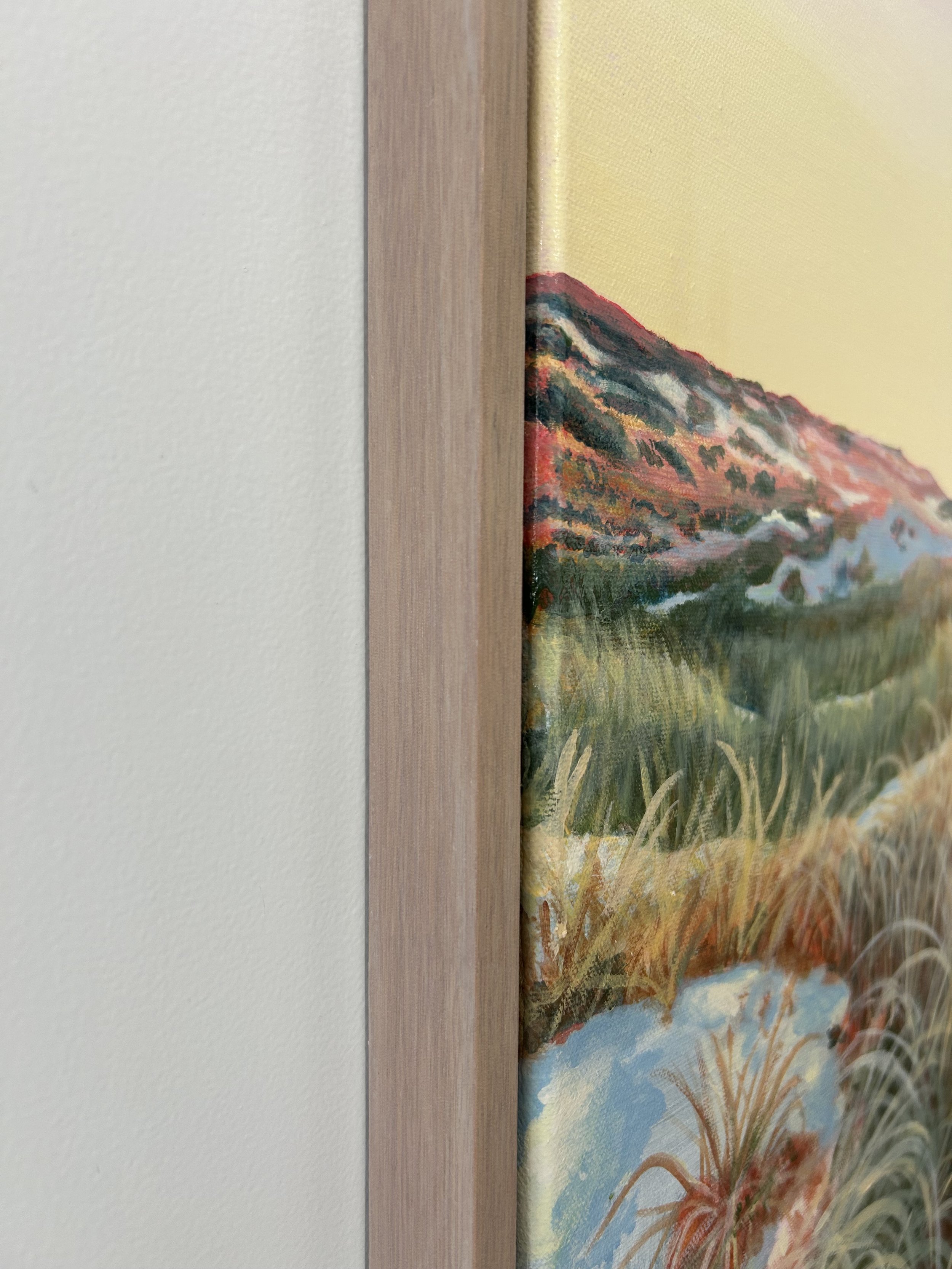 Close-up of a painting featuring a landscape with mountains, greenery, and water, framed with a light hardwood. The painting features a soft pink sky with peachy coloured sand dunes.
