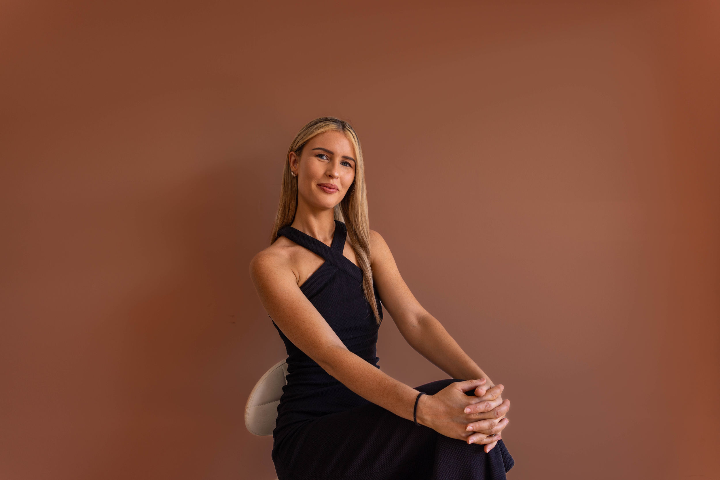 A woman with long blonde hair sitting on a white chair against a plain brown background, wearing a black sleeveless dress, looking forward.