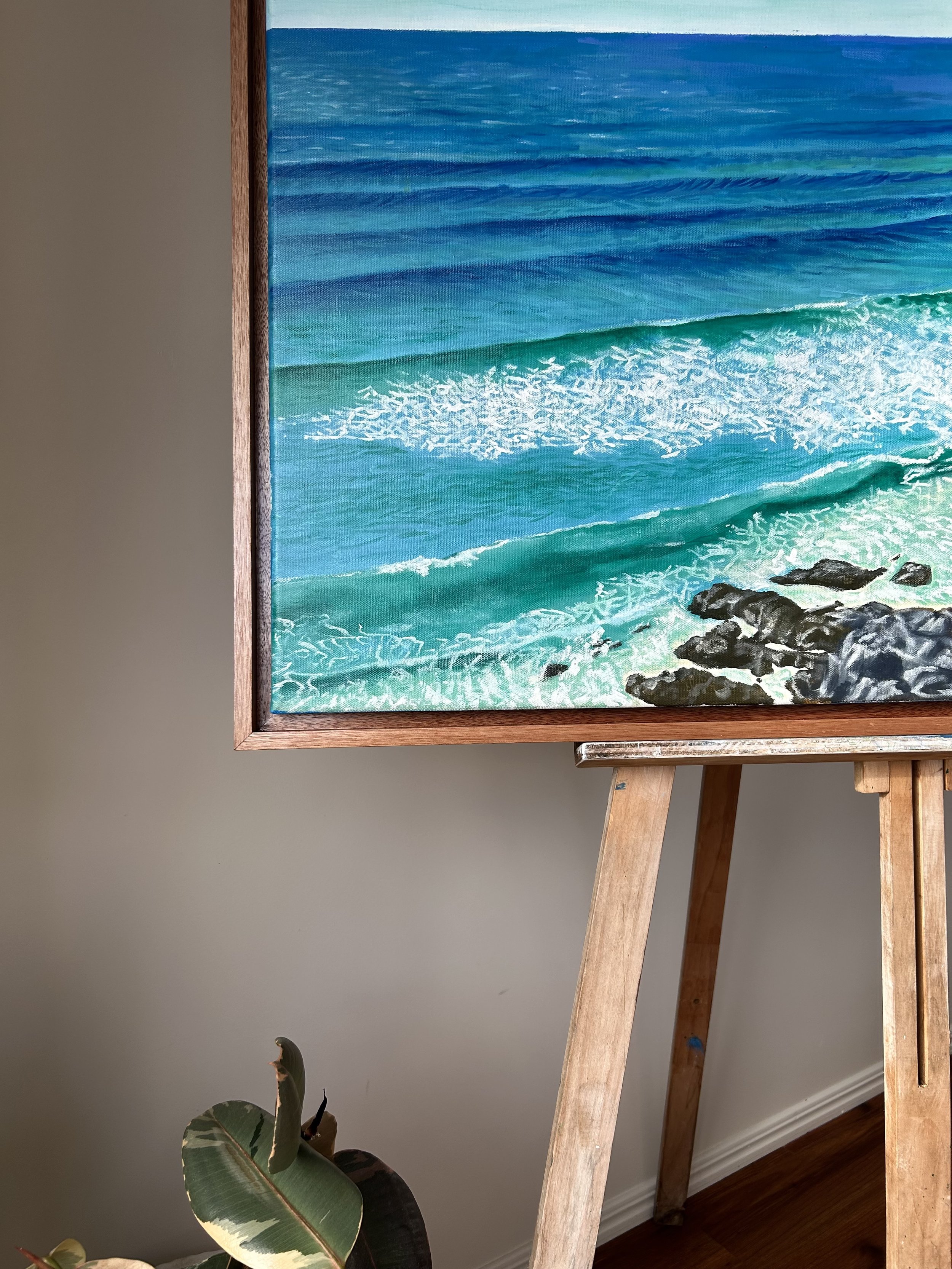 A painting of Coolum Beach showing a blue ocean with waves crashing on rocks, framed and displayed on a wooden easel indoors. A potted plant is partially visible in the foreground.