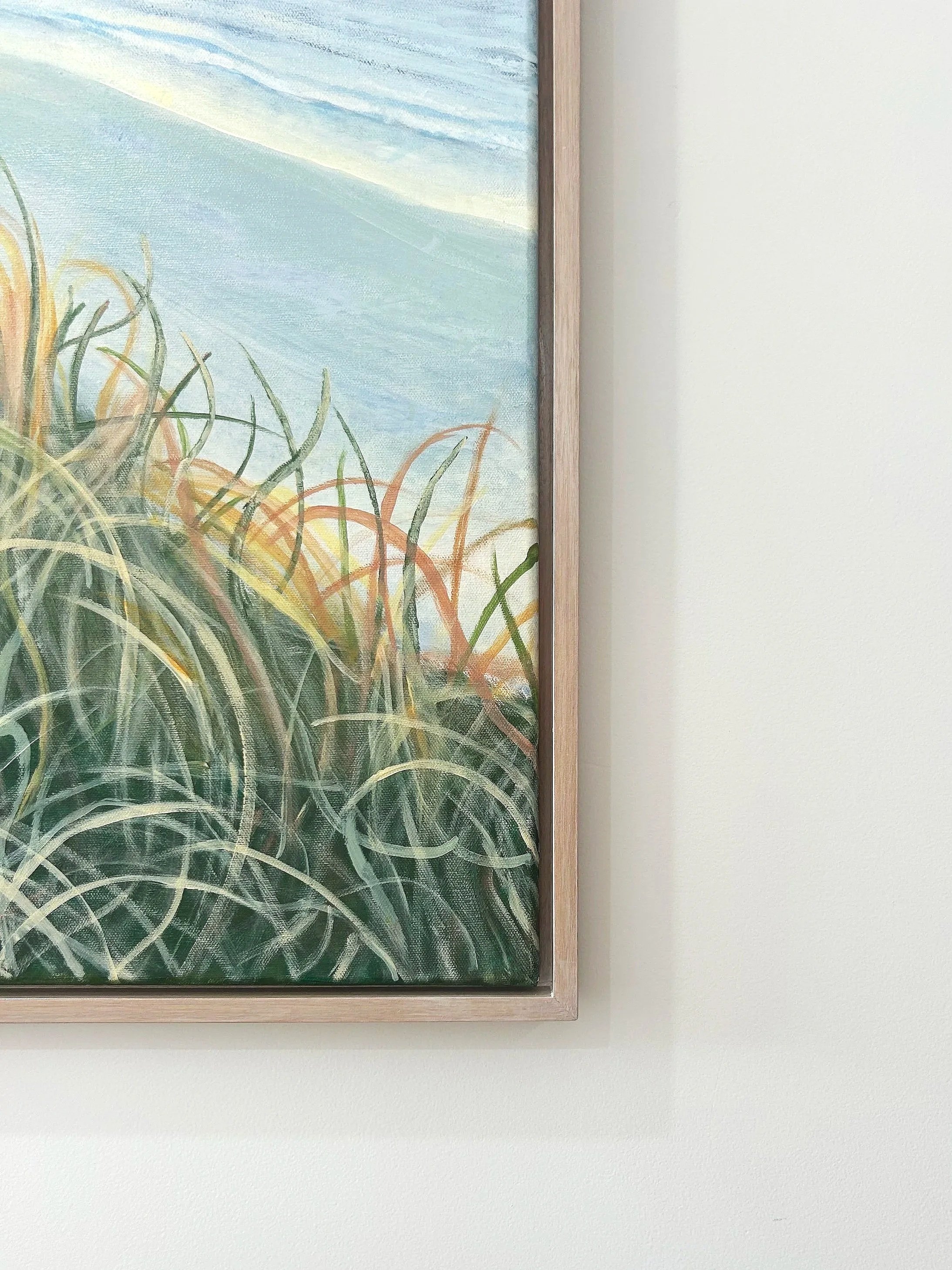 Part of a framed painting of beach grass, with sand and sky in the background, mounted on a white wall.
