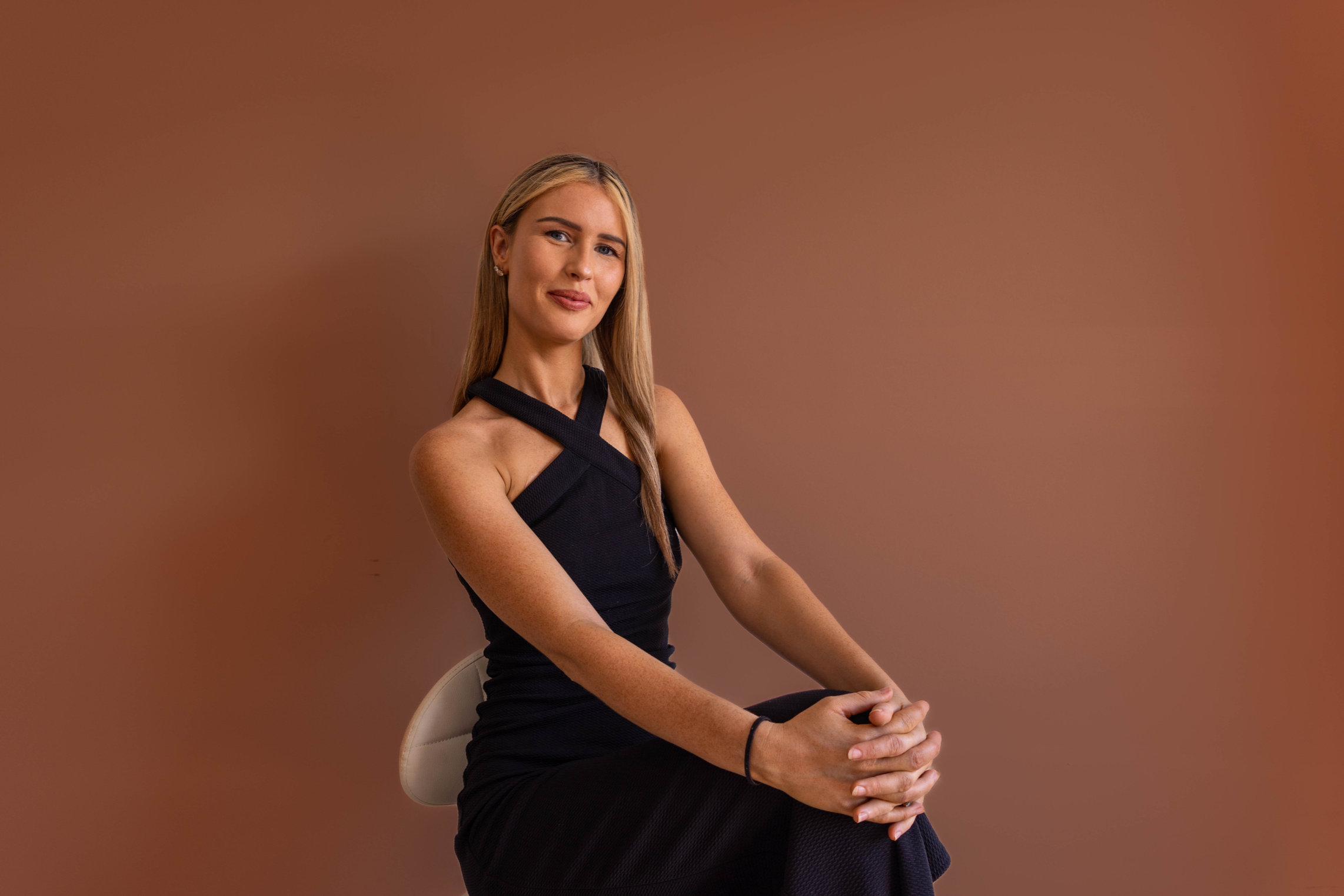 A woman with long blonde hair wearing a black sleeveless dress sitting on a beige chair against a brown background, looking forward.