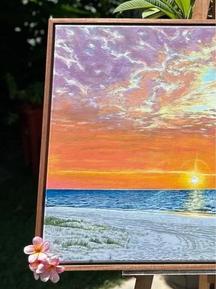 Painting of a sunset over the ocean, with a orange and pink sky, and a few clouds. The sun is near the horizon, reflecting on the water. The sandy beach in the foreground has some green grass patches, and there is a pink flower attached to the bottom left corner of the frame.