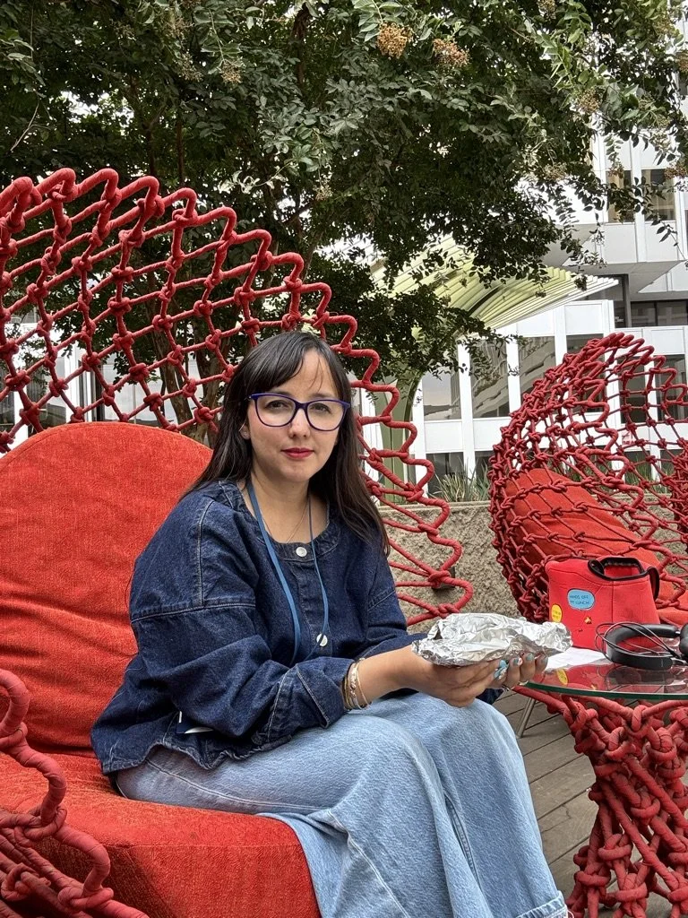 A woman with dark hair and glasses, wearing a denim jacket, sitting on a red outdoor chair holding a foil-wrapped food item, in a garden setting with trees and modern buildings in the background.
