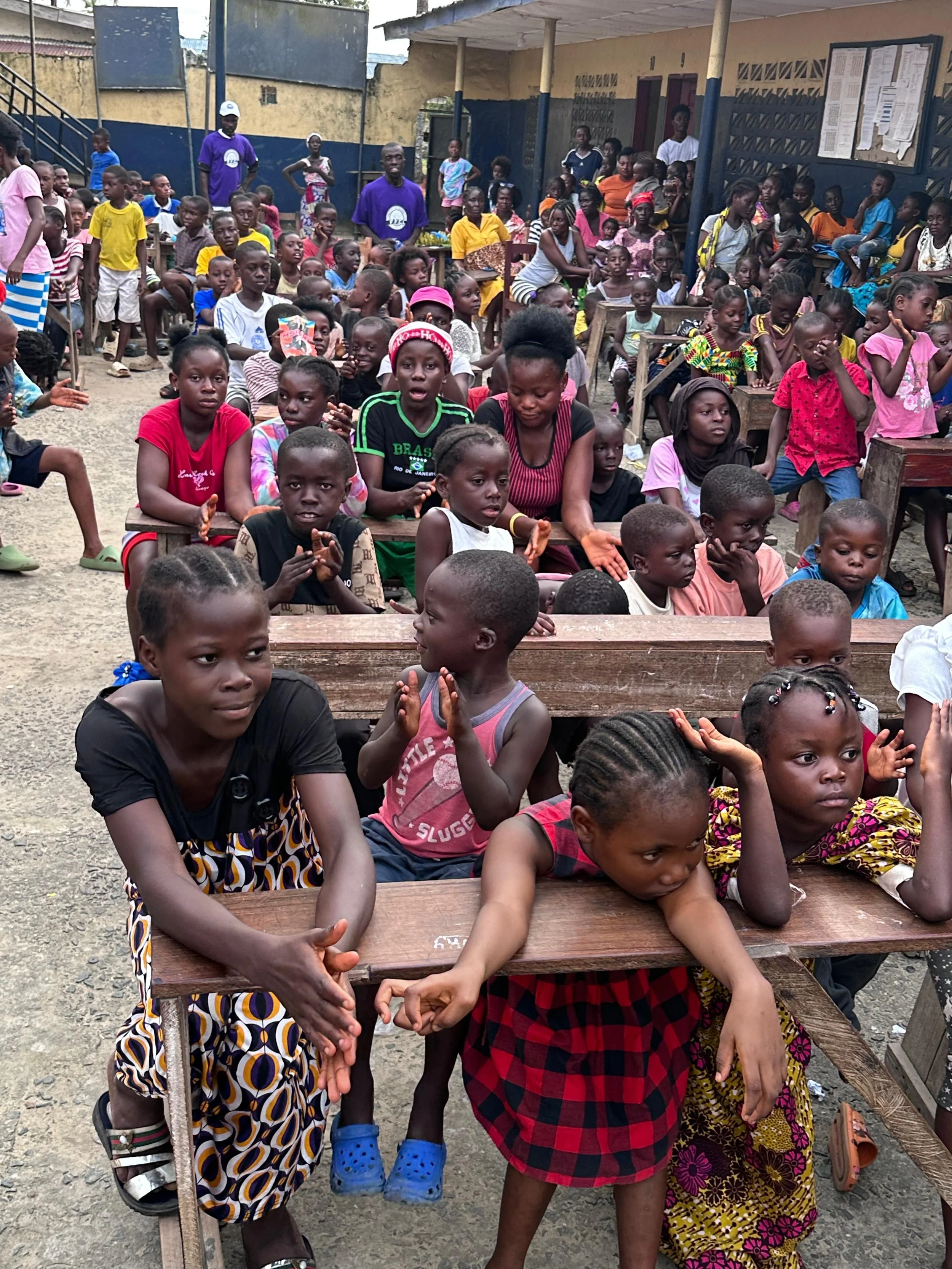 Children and adults gathered in an outdoor schoolyard. Some children sit at wooden desks while others are standing or sitting on benches. The background shows a building with notice boards and some adults supervising.