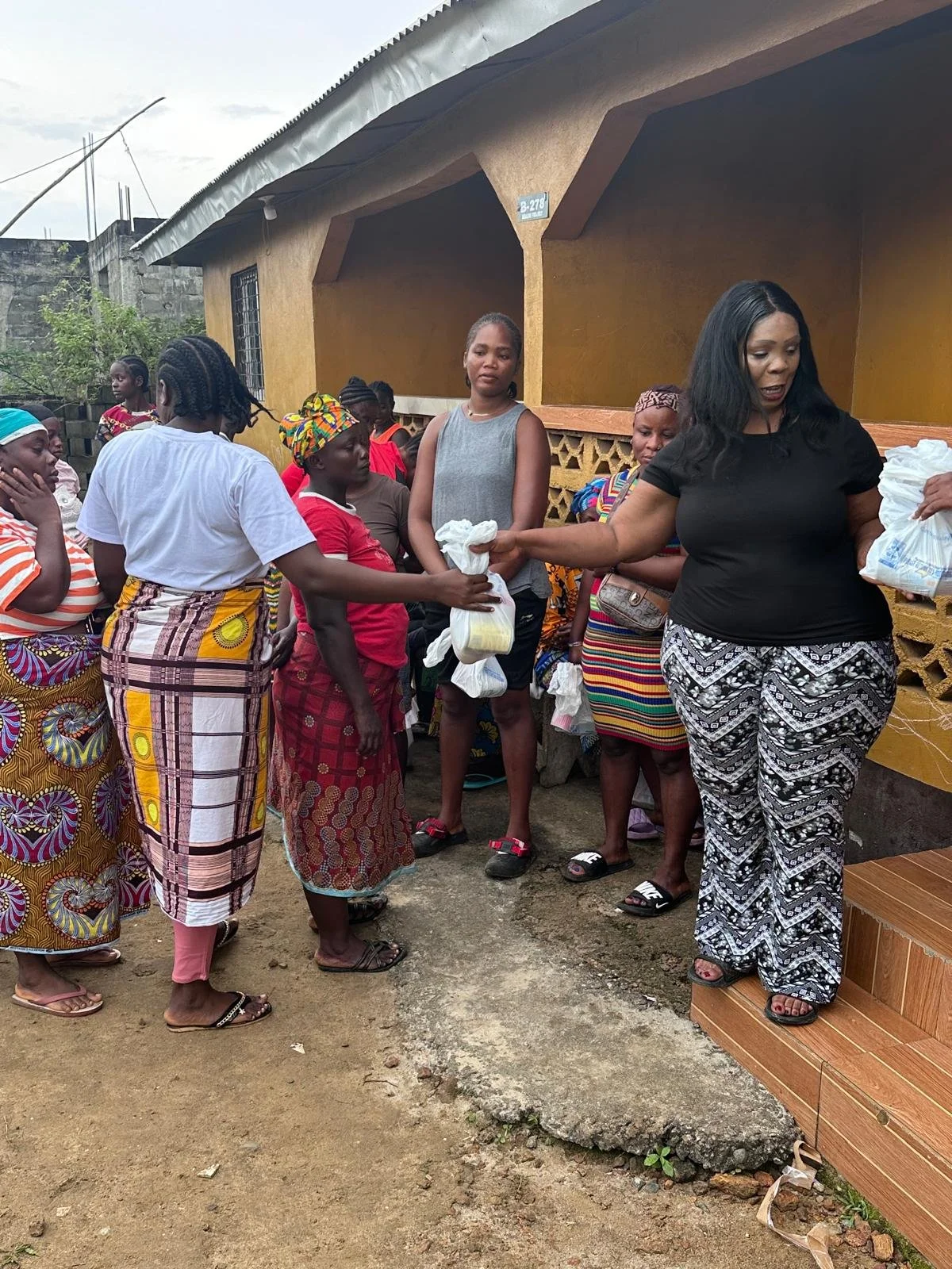 A group of women and girls standing outside a yellow building, receiving food or supplies from a woman standing on a small porch.
