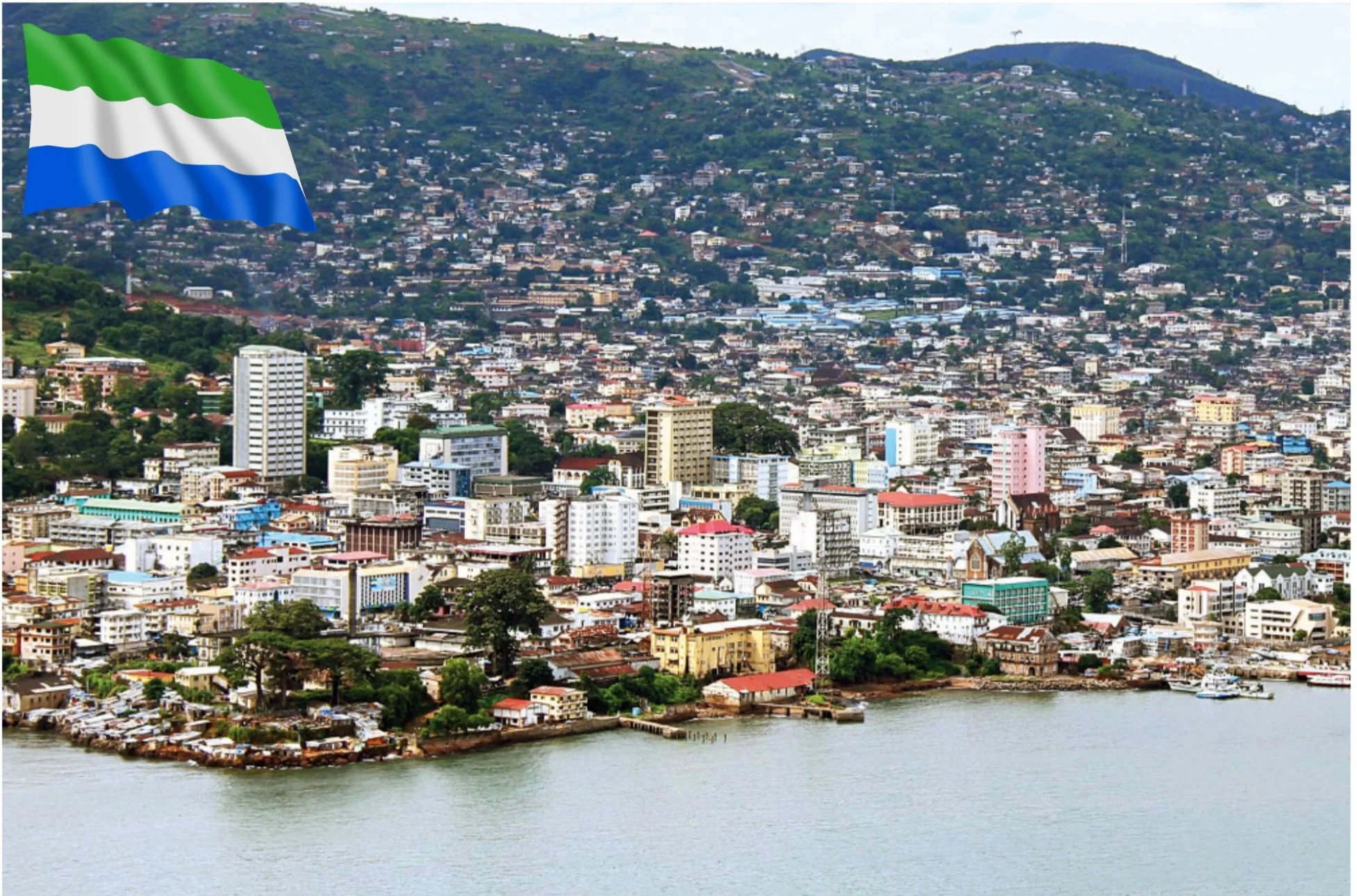 A cityscape of San Francisco with various buildings along the waterfront, hills in the background, and a large flag with green, white, and blue stripes in the top left corner.