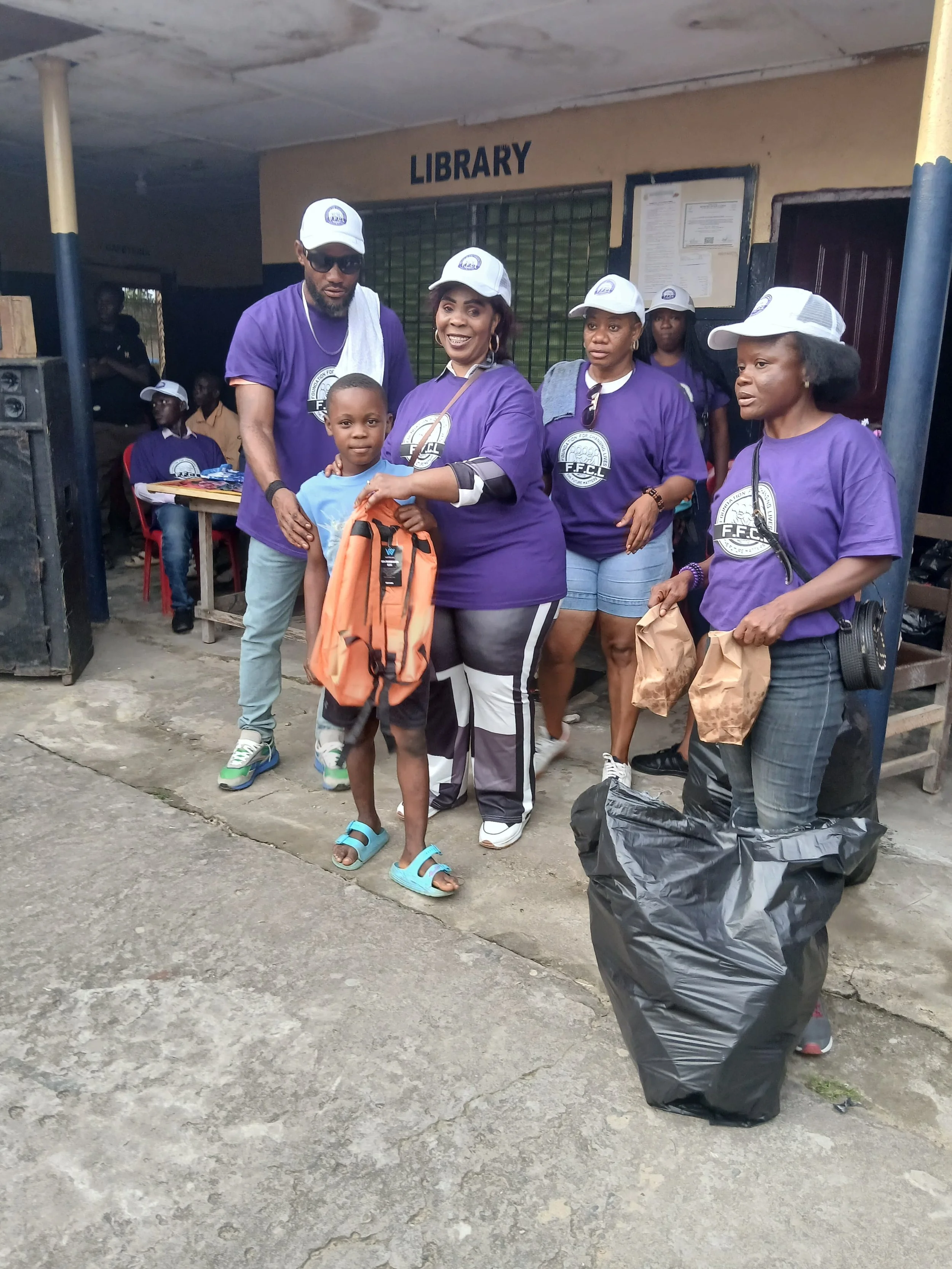 Group of people, including a boy with an orange backpack, standing outside a building labeled 'Library'. Most are wearing purple shirts and white hats, and there are bags and supplies in the foreground.