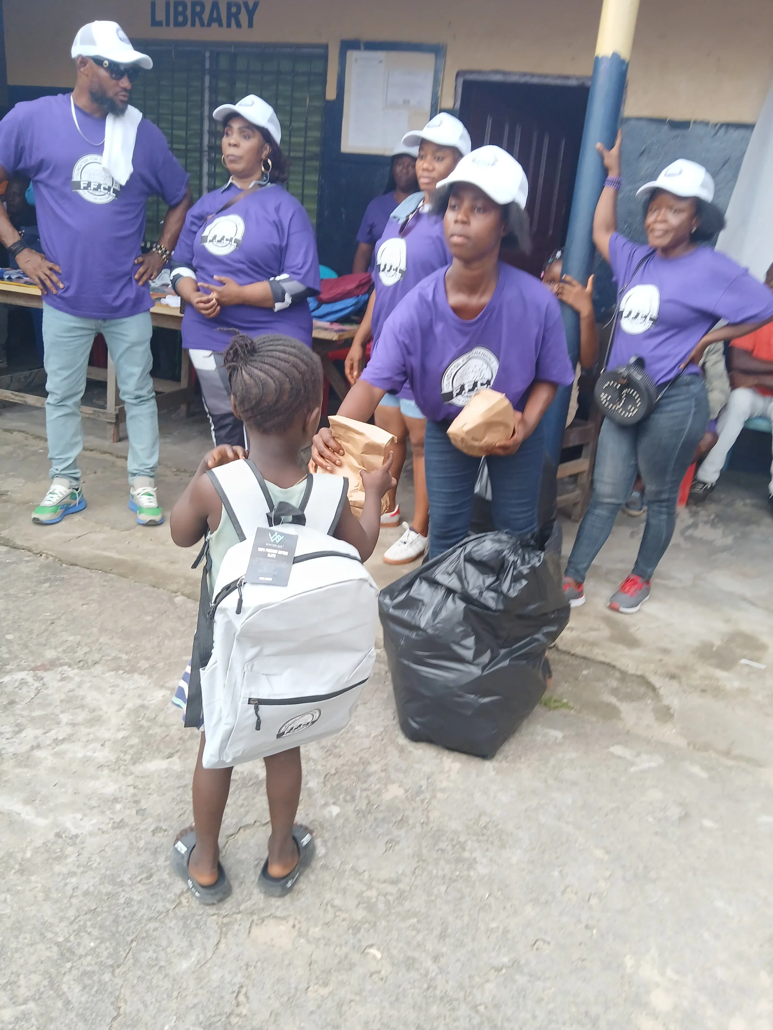 A young girl with braided hair and a white backpack exchanging a bag of food with an adult woman wearing a purple t-shirt, standing in front of a group of people in purple t-shirts and white hats, outside a building with a sign that says