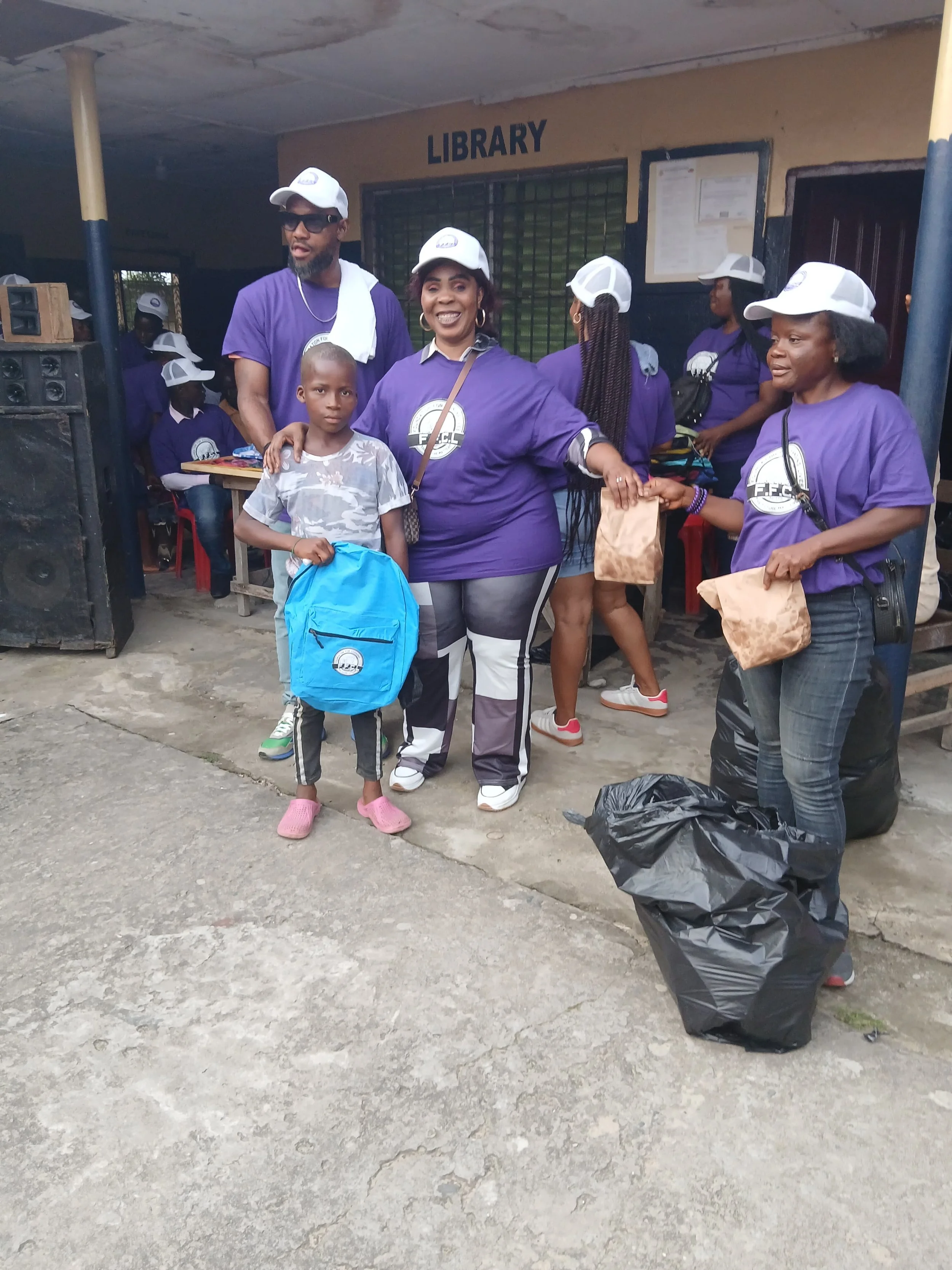 Group of people standing outside a library, wearing matching purple shirts and white caps, with some holding brown paper bags and a child holding a blue backpack.