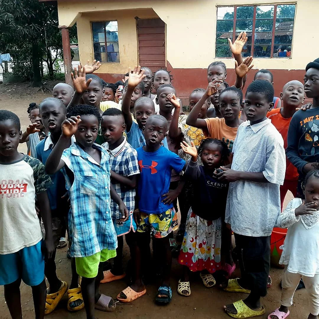A group of children, some raising their hands, stands outside a building with windows. They appear to be in a schoolyard or community area, surrounded by trees and a dirt ground.