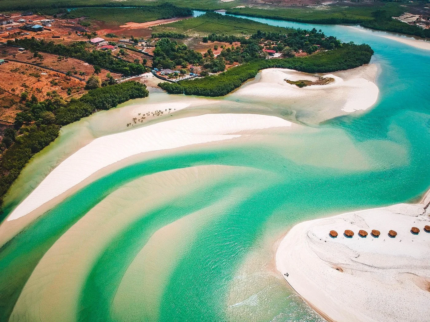 Aerial view of a river with turquoise water flowing through white sandy beaches and lush green vegetation, with some small structures and trees along the riverbank.