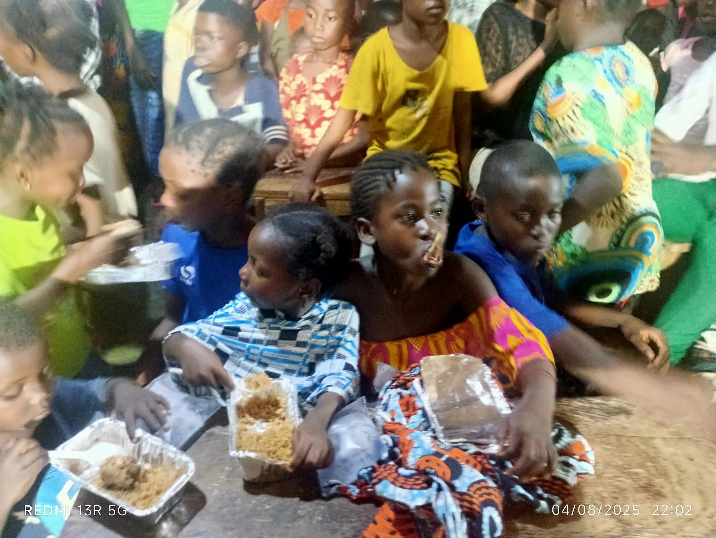 Group of children gathered closely together, some sitting and others standing, with a woman in colorful traditional attire seated in front. The children are holding or reaching for food containers of rice. The background shows a crowded setting with more children and adults.
