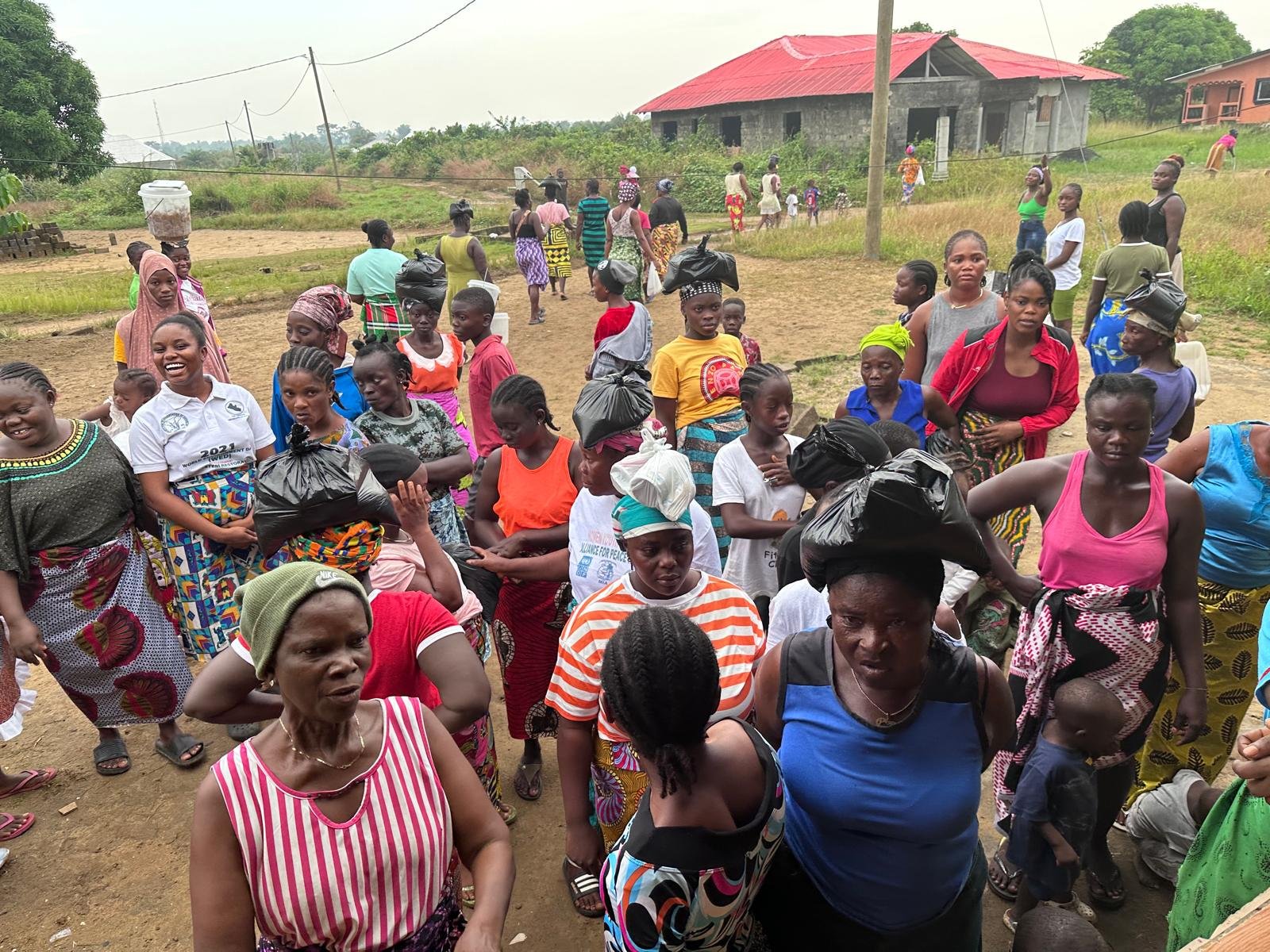 A large group of women and children gathered outdoors in a rural area, some carrying black plastic bags on their heads. The background shows a partly constructed building, grassy fields, and people walking around.