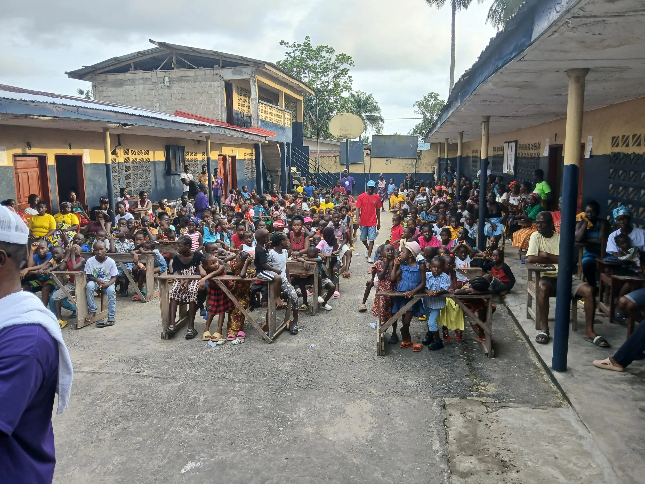 A large group of children and adults gathered in a school courtyard surrounded by classrooms. Some children are sitting at desks, others are standing or sitting on the ground. Adults are standing or sitting along the sides, and the background shows school buildings and a basketball hoop.