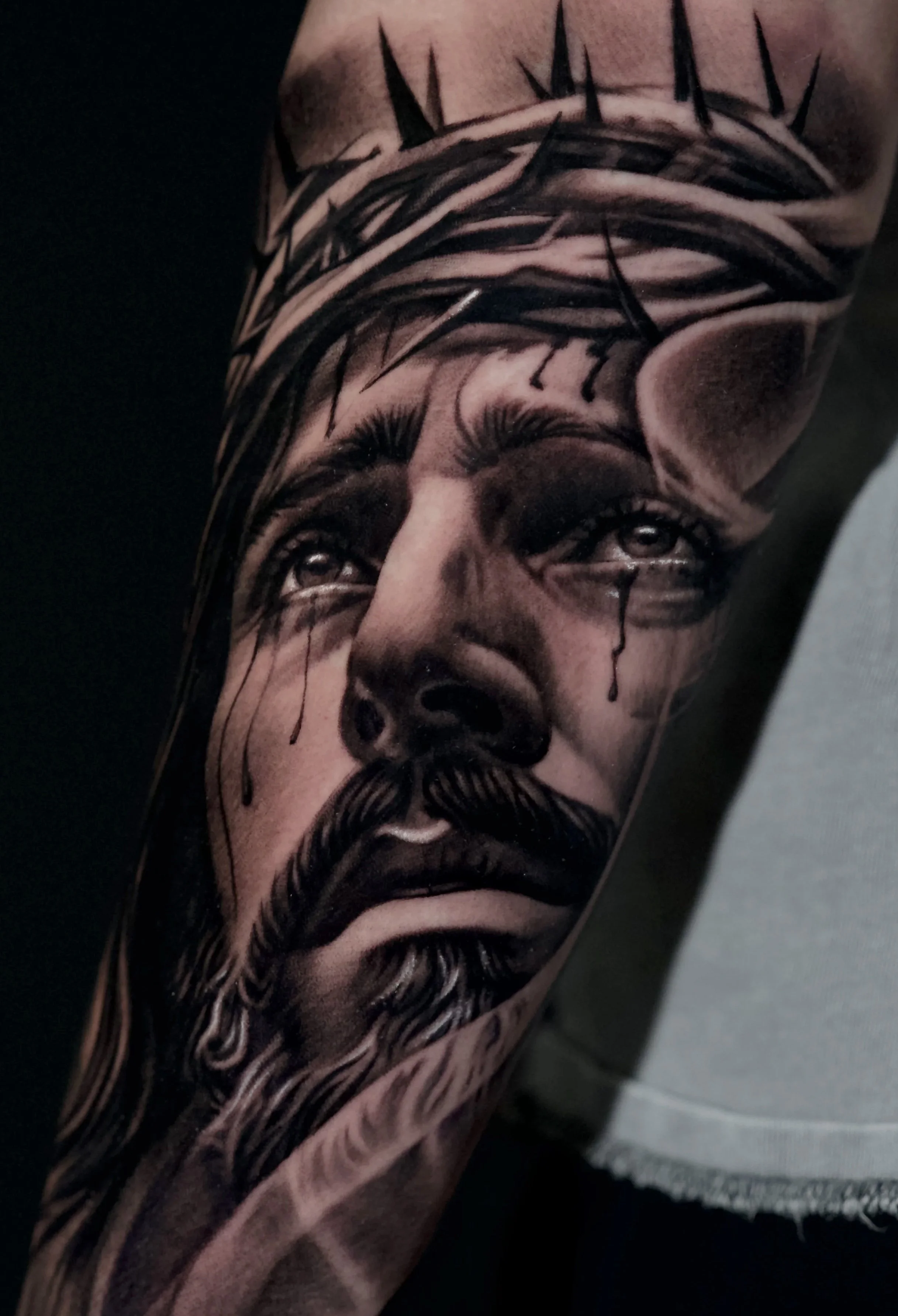 Close-up of a tattooed person's arm, featuring a detailed black-and-gray portrait of a Jesus Christ. Tattoo done by Allan Mandi realism tattoo Artist from Knoxville Tennessee