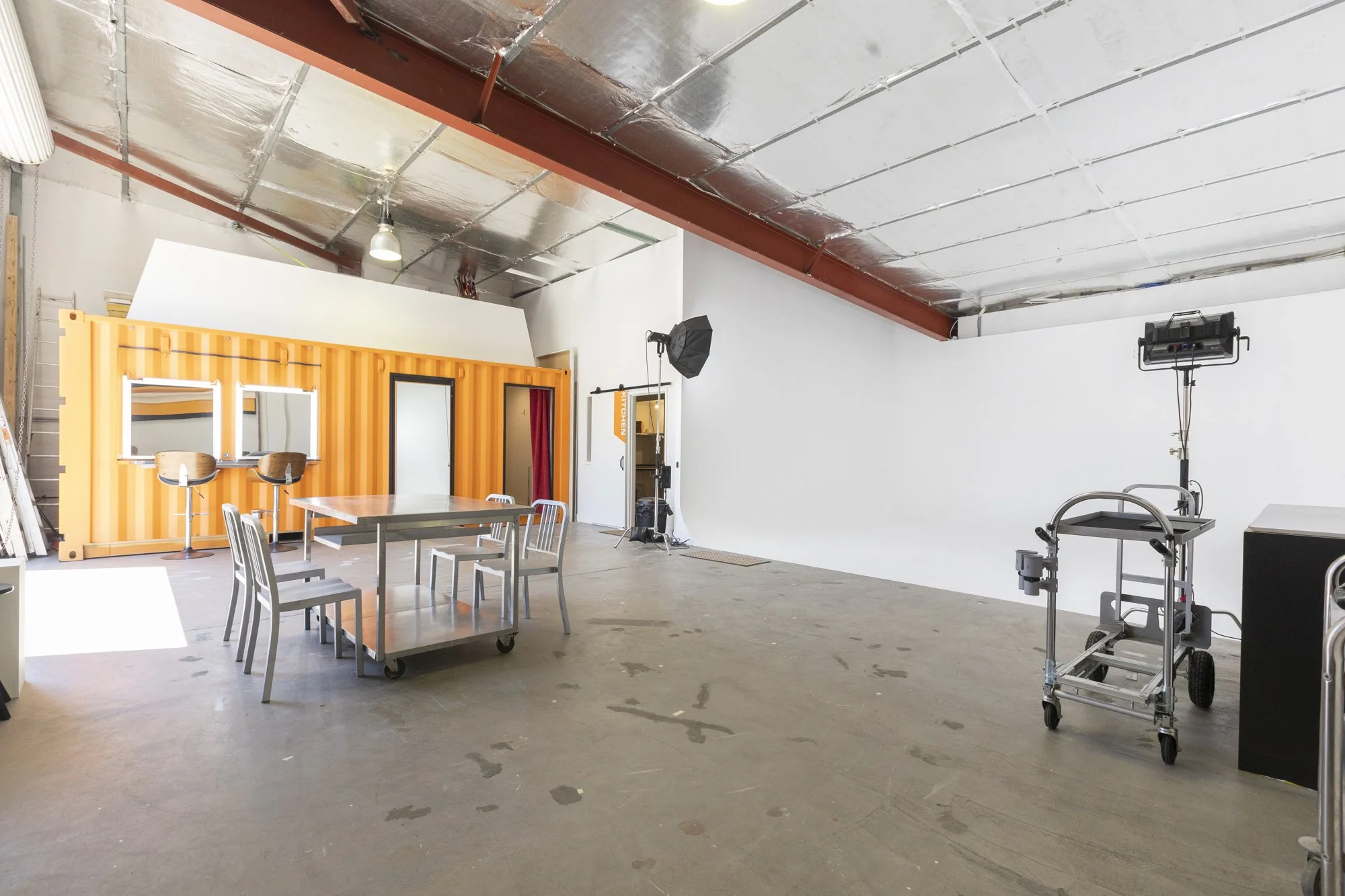 Interior of a photography studio or film set with professional lighting equipment, a white backdrop wall, a small table and chairs, and a large shipping container painted orange on one side.
