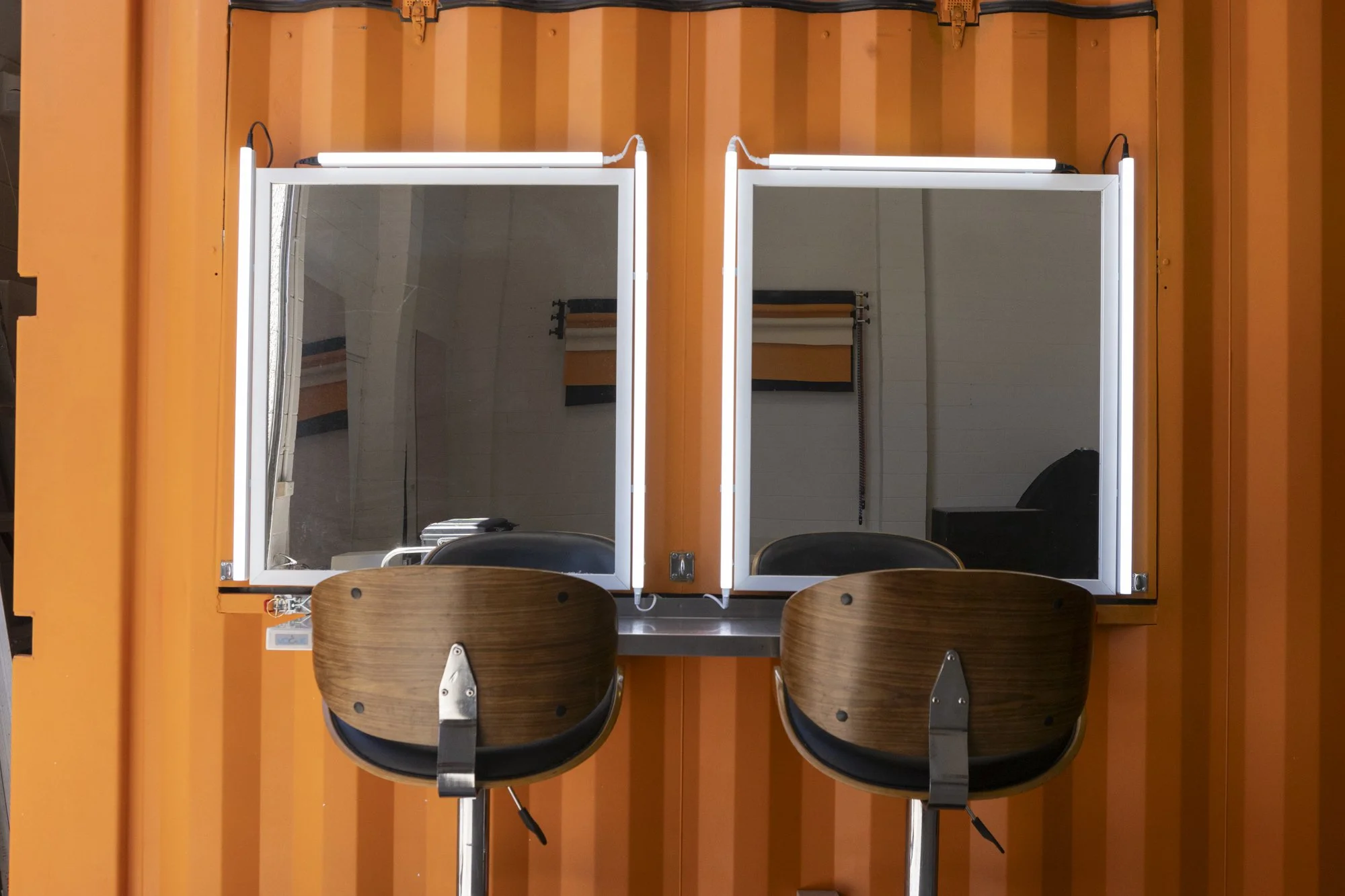 Two wooden salon chairs in front of a large backlit mirror with two sections, mounted on an orange wall. The mirror reflects an interior space with white walls and a black object, possibly a cabinet or equipment.