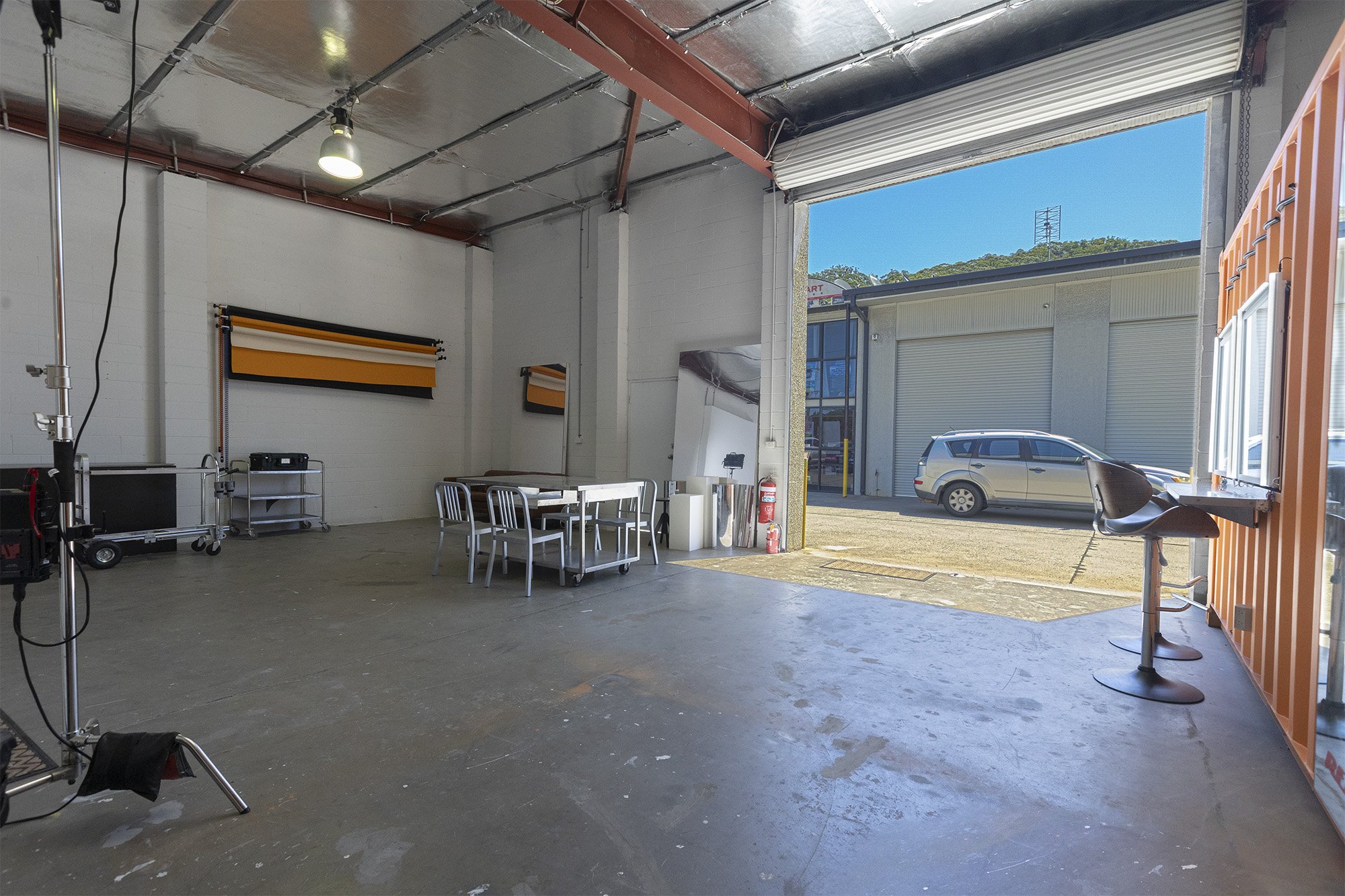 Empty industrial garage or studio space with a large open rolling door, a white table and chairs, some equipment and a high chair, with a view of parked cars outside under a blue sky.