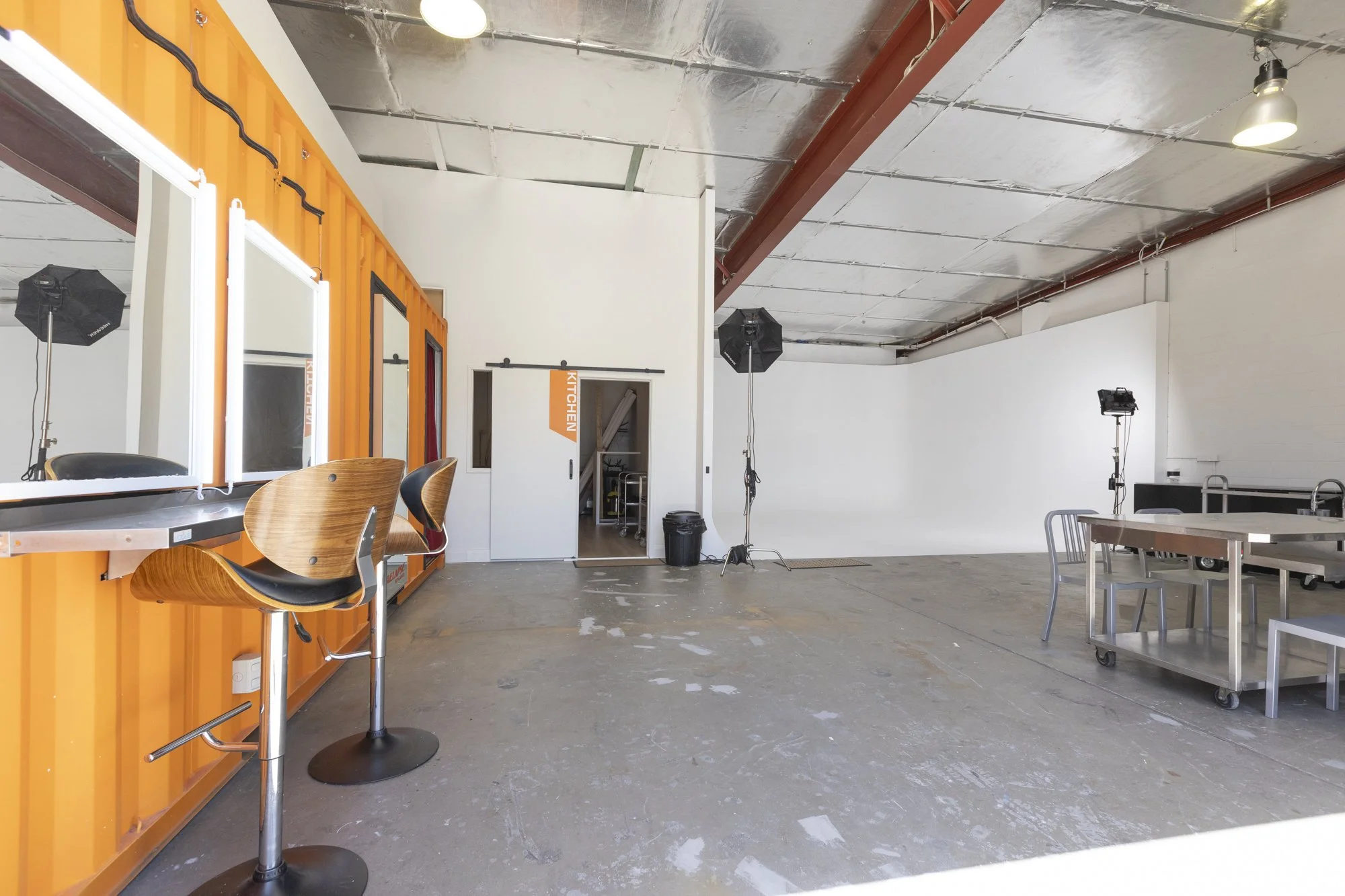 Empty photography or film studio with a white brick wall, a bright orange wall with mirrors, a couple of studio lights and chairs, and a door labeled 'KITCHEN.'