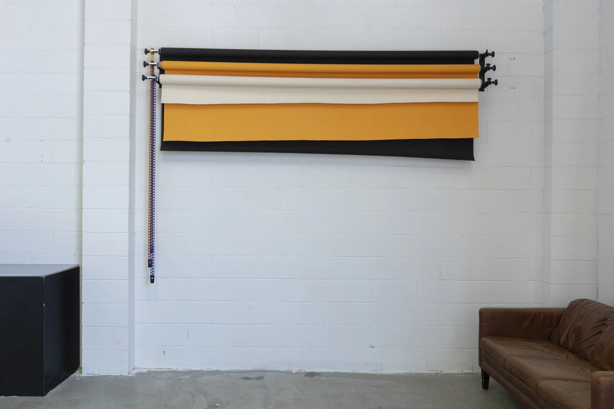 Studio photo backdrop with black, white, and yellow fabric rolls mounted on a wall, and a brown leather sofa in a minimalist room with white brick walls.