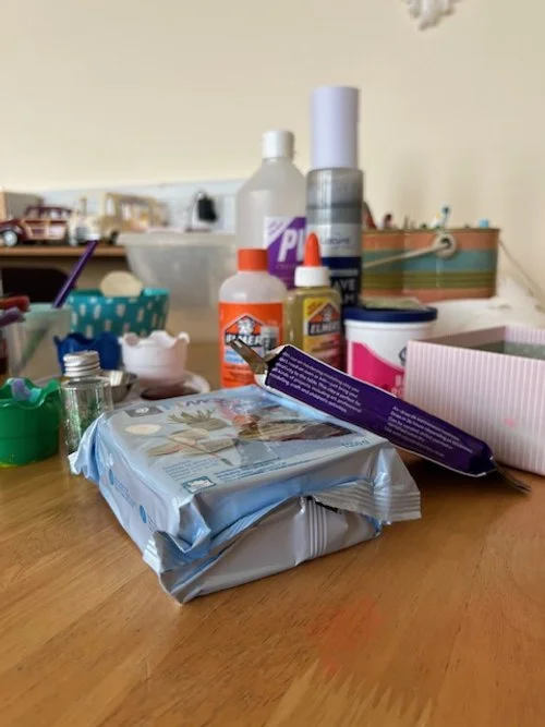 A cluttered tabletop with household items including a pack of baby wipes, glue bottles, a roll of tape, a small container, a spray can, and various craft supplies.