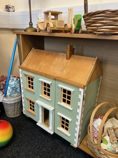 A large wooden dollhouse with a green exterior, brown roof, and white and brown window and door frames, positioned under a wooden table with toys on top, including a wooden truck and lamp.