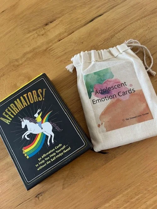 A box of Aftermaters! emotion cards and a cloth bag labeled "Adolescent Emotion Cards" on a wooden surface.