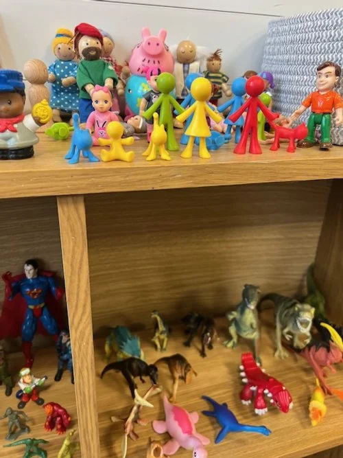Toy figurines arranged on a wooden shelf, including Peppa Pig, children figures in various colors, a Superman figure, horses, and dinosaur toys.