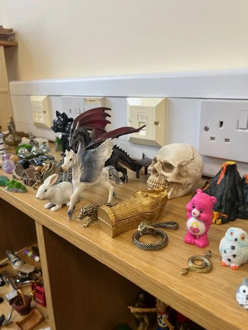 Collection of toy figures and objects on a wooden shelf, including dragons, animals, skull, pink bear, and various other toys.