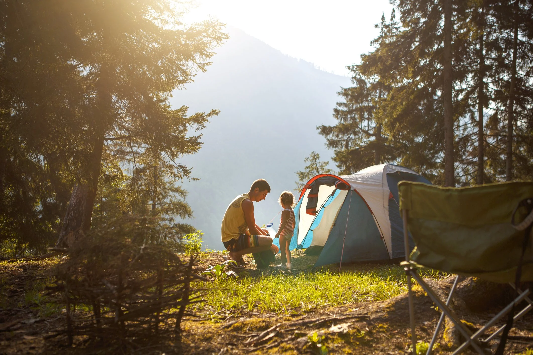A man and a young girl standing near a tent in a forest, with mountains in the background, during sunset.