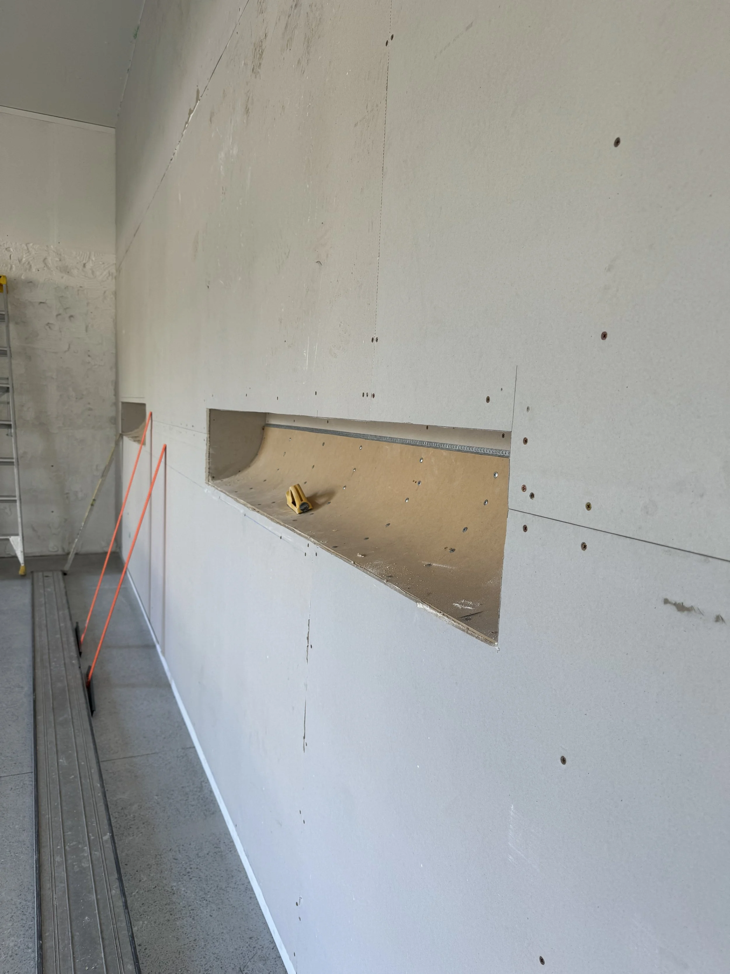 Interior wall under construction with an opening and drywall panels, with tools and equipment visible.