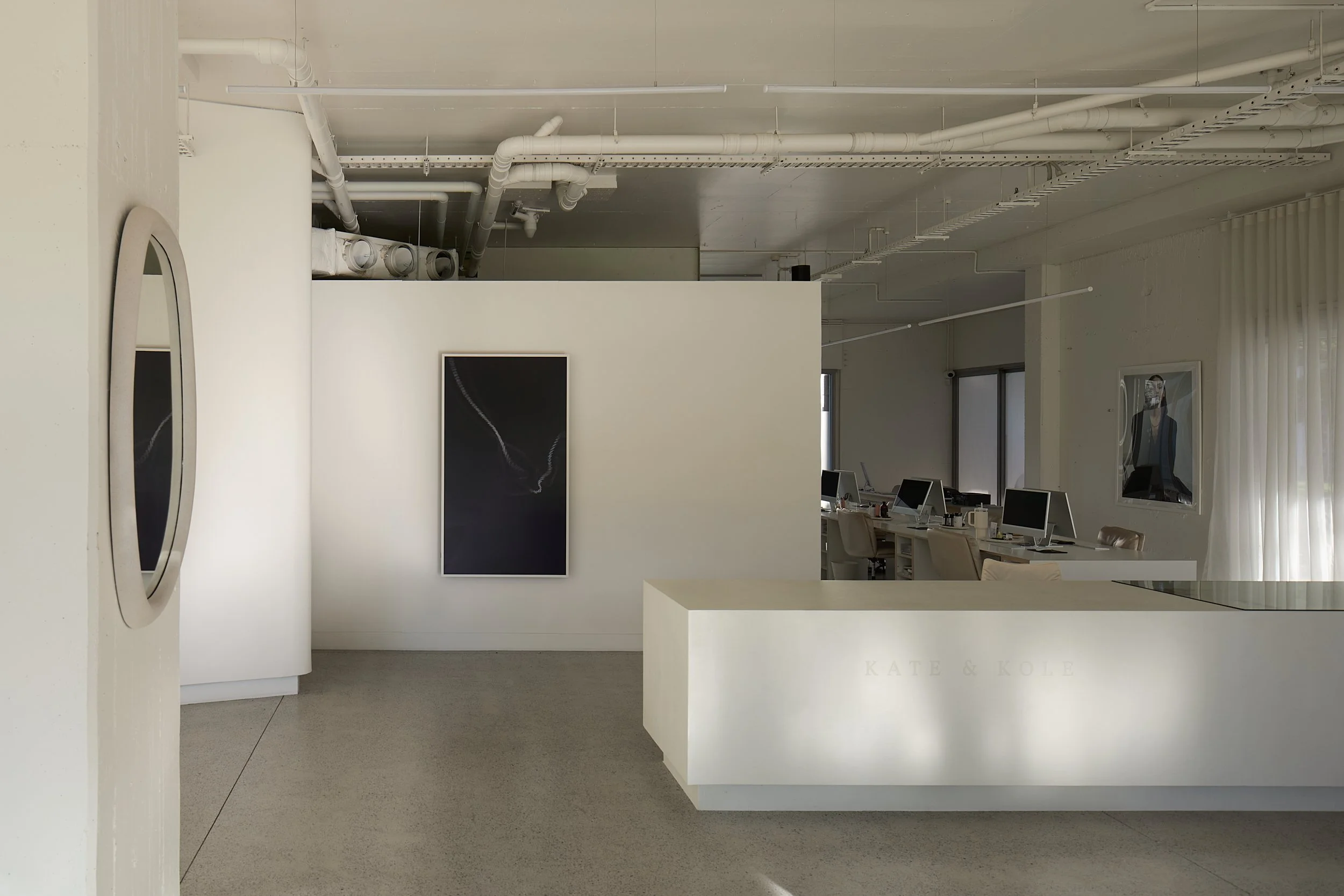 Interior of a modern art gallery with white walls, black artwork, computer desks, and curtains on windows.