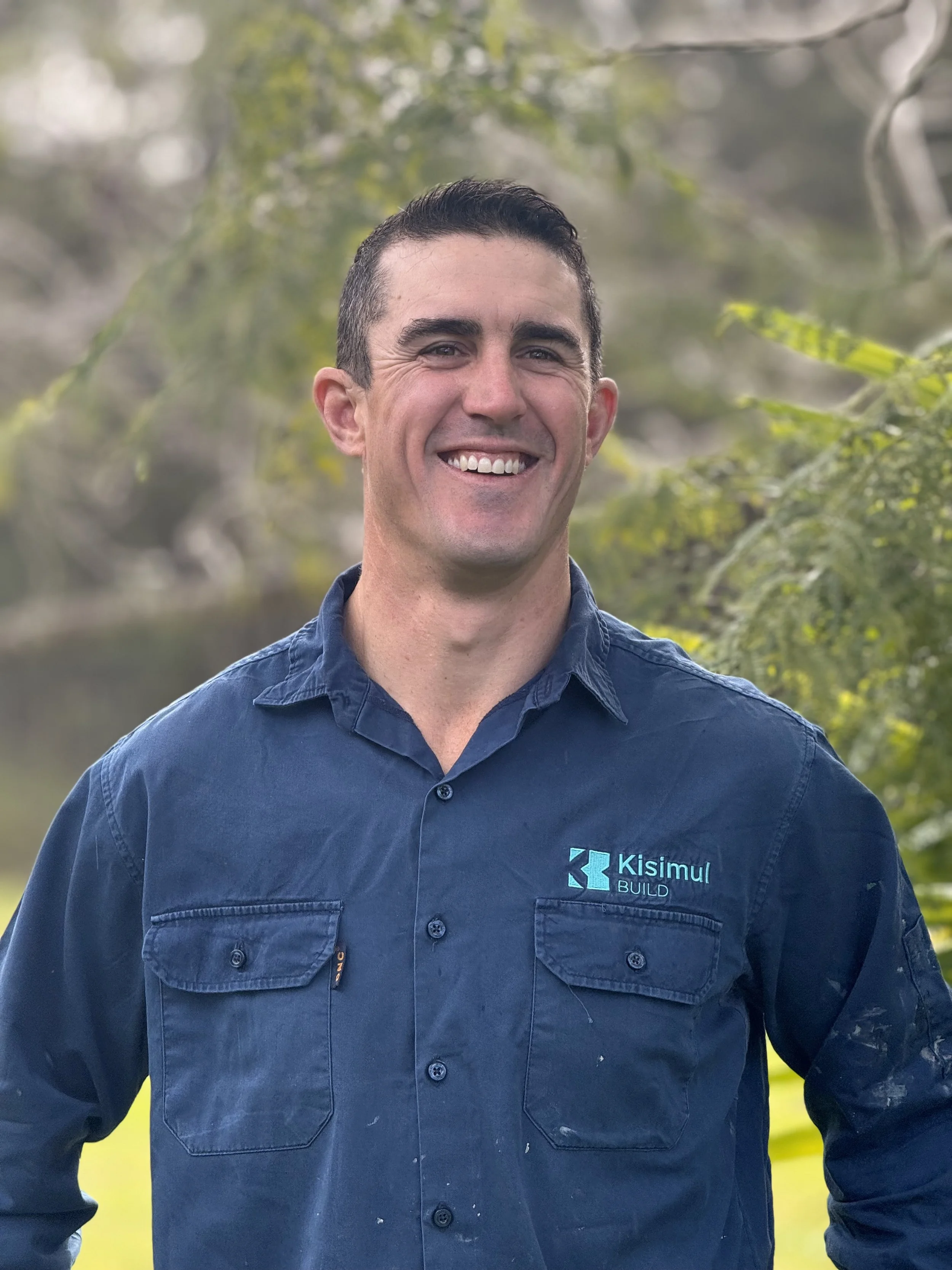 A smiling man with dark hair, wearing a navy blue work shirt with the logo 'Kisimul BUILD,' standing outdoors with blurred trees and water in the background.