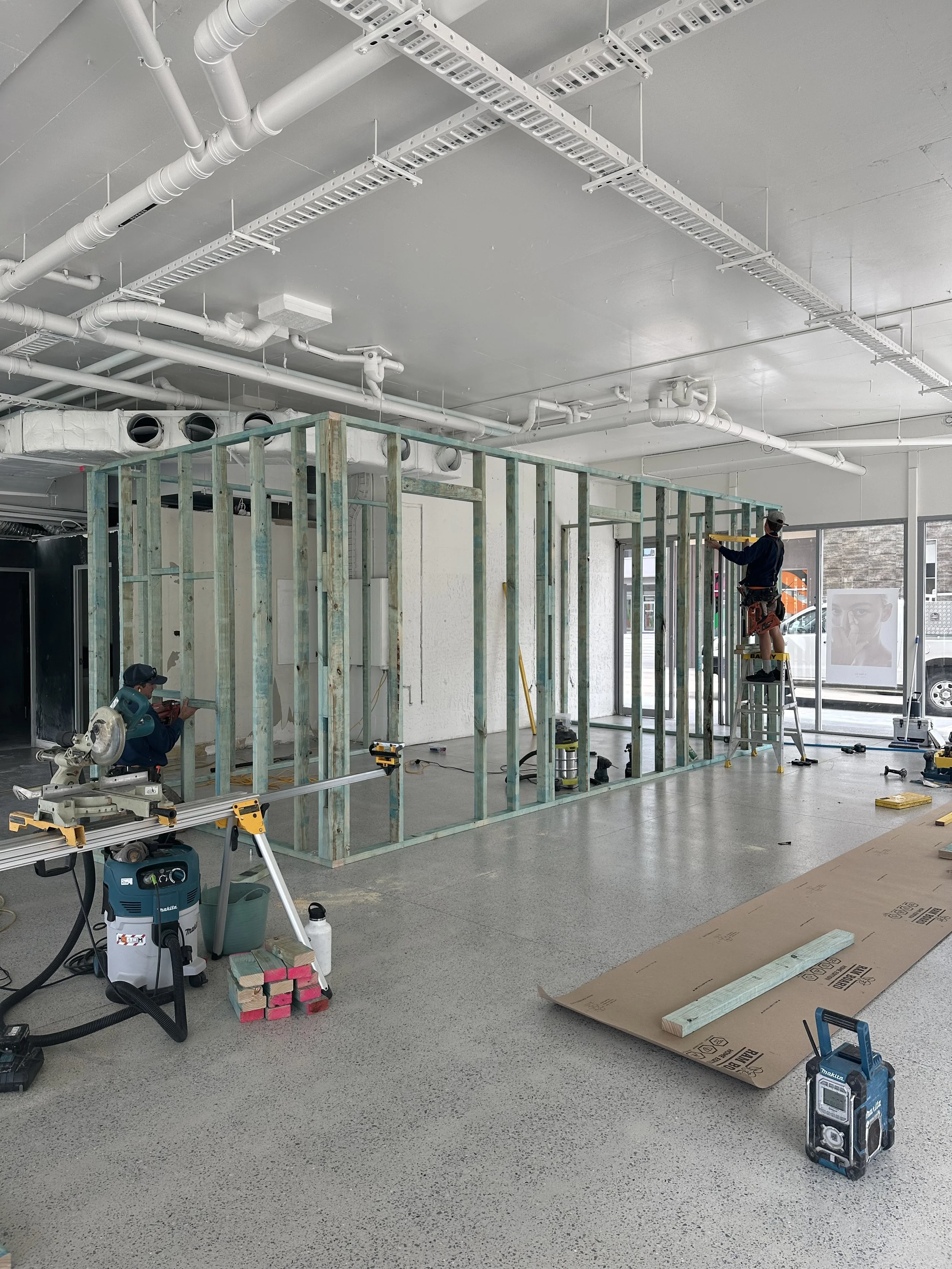 Construction workers building a wall inside a commercial space, with visible framing, tools, and supplies on the floor.