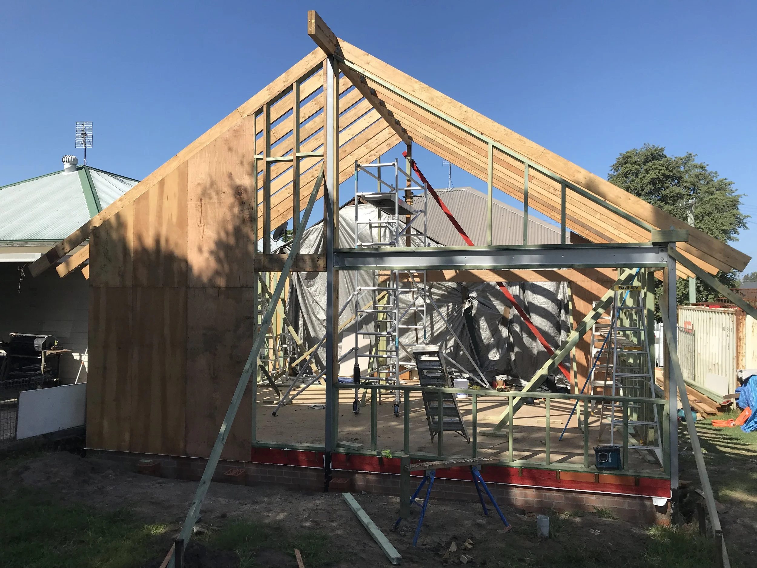 A house under construction with wooden framing and metal supports, surrounded by construction tools and scaffolding, in a backyard with a neighboring house with a green roof.