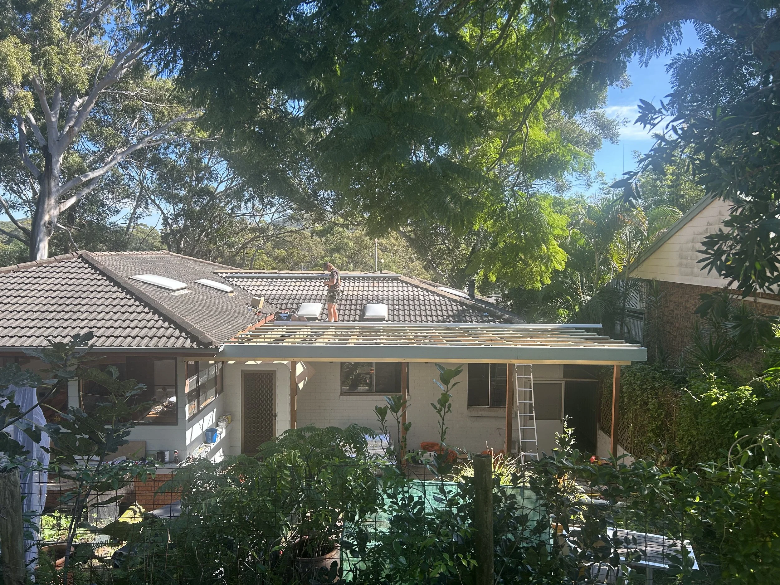 Construction worker on house roof installing solar panels, ladder nearby, surrounded by trees and neighboring houses.