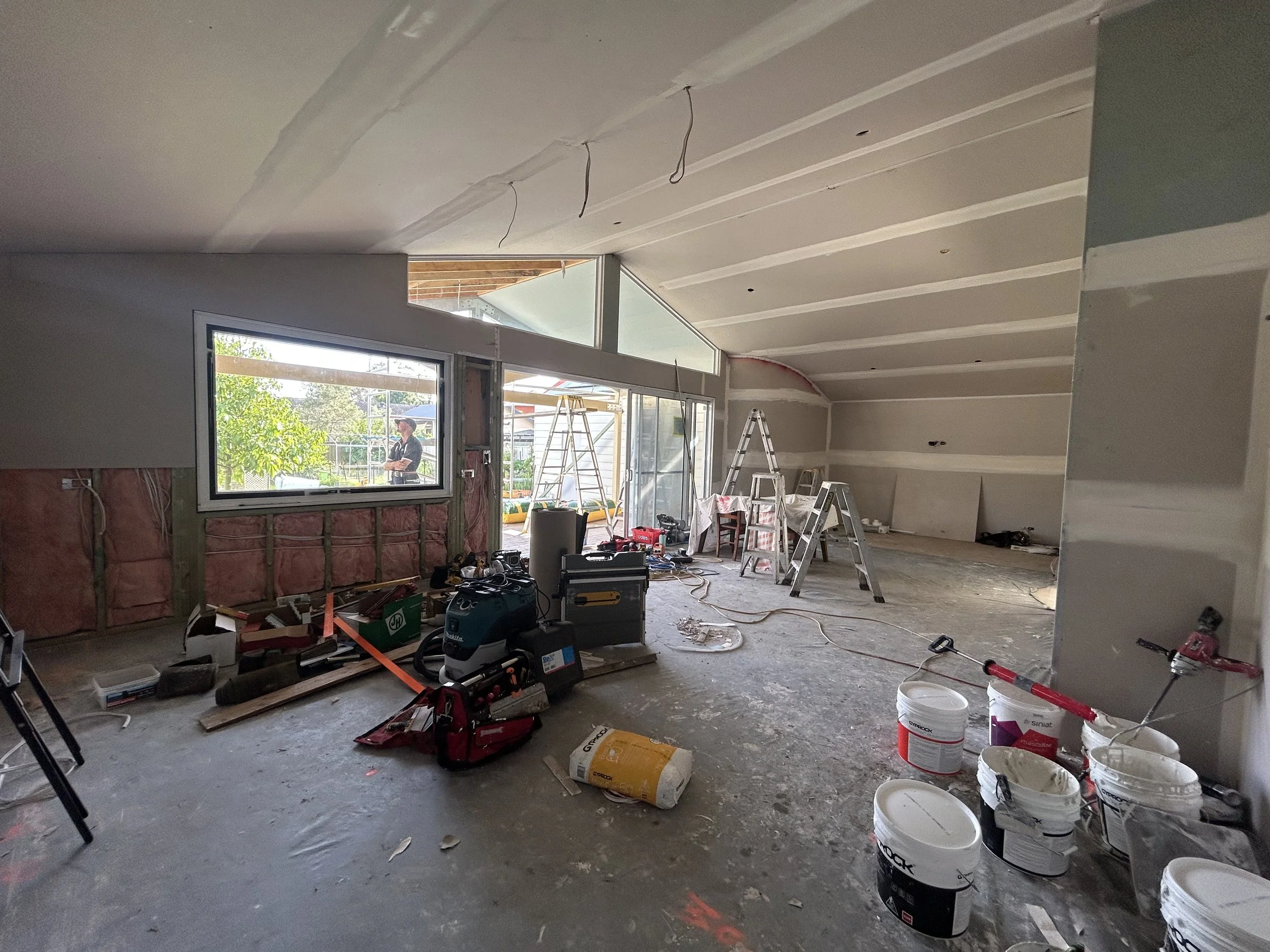 Interior of a house under construction with tools, ladders, buckets, and materials scattered on the floor, and a worker visible outside through the window.