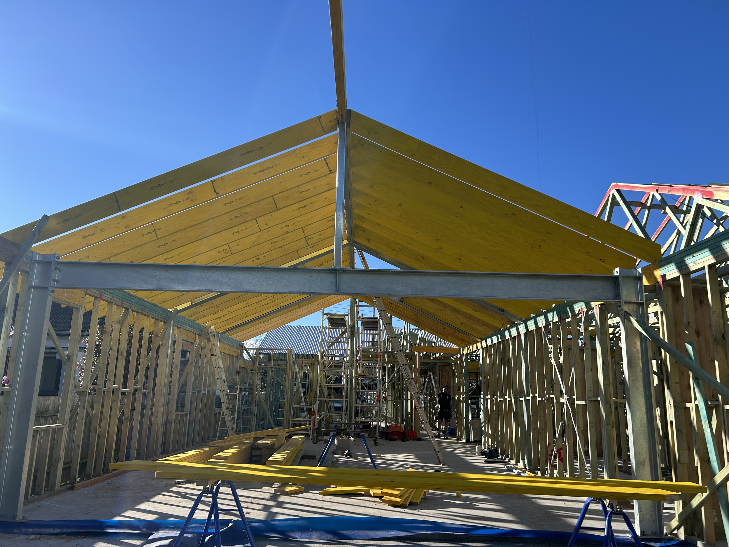 Construction site with wooden framing and yellow roof panels being assembled under clear blue sky.