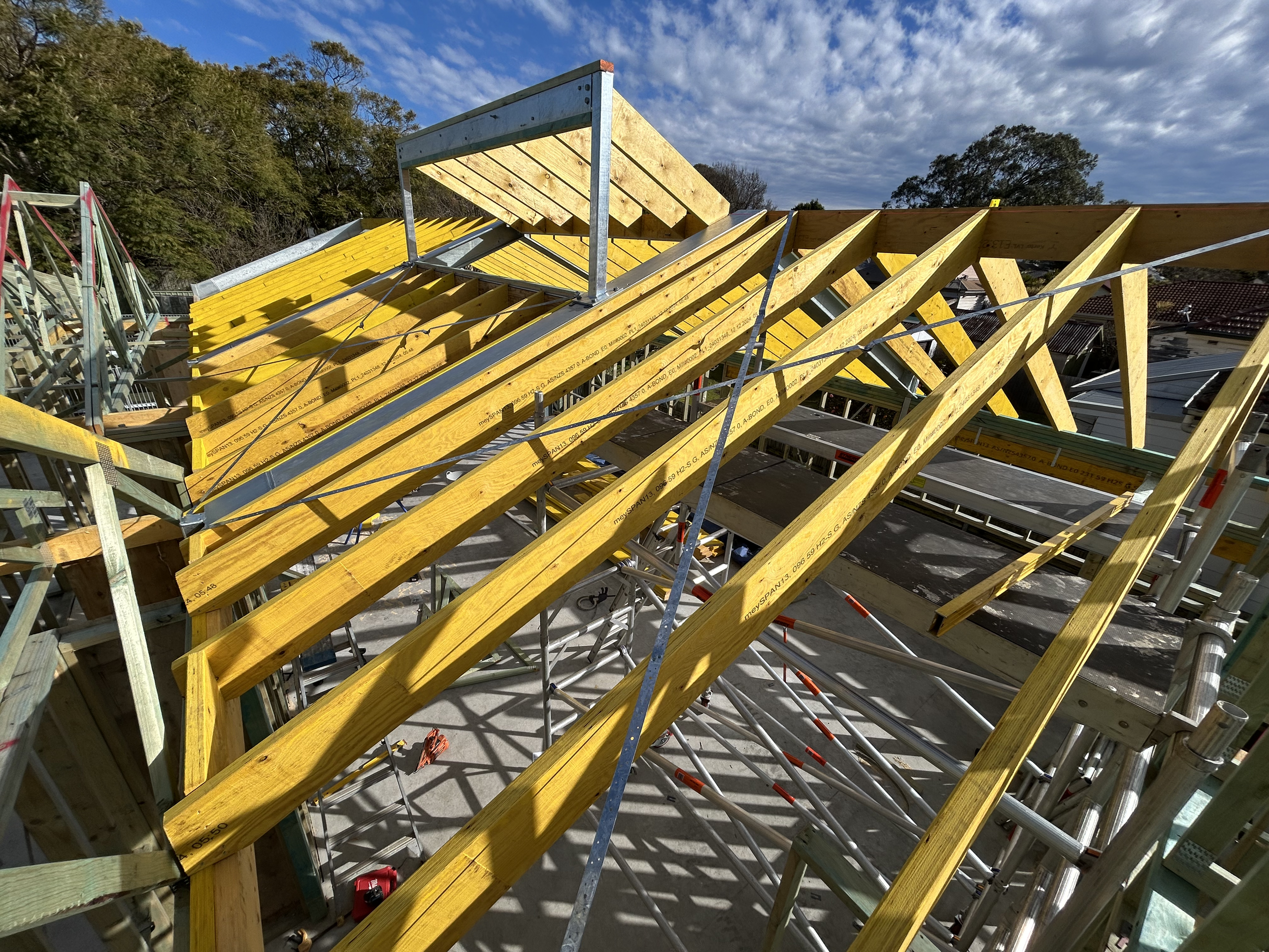 Construction site with wooden roof rafters and scaffolding under a partly cloudy sky.