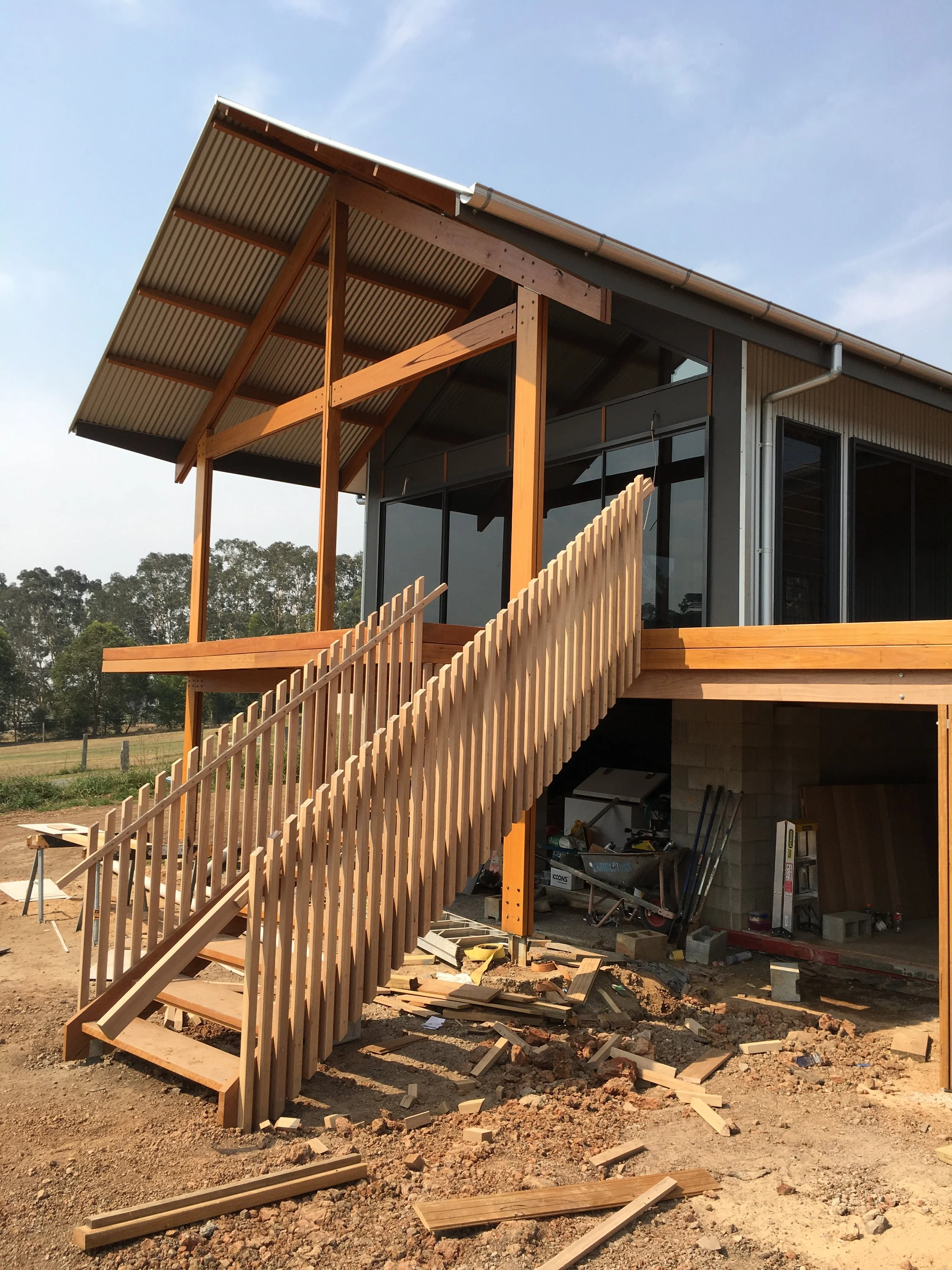House under construction with wooden staircase leading to a second-story deck, construction materials and tools underneath, and a partially completed roof and exterior walls.