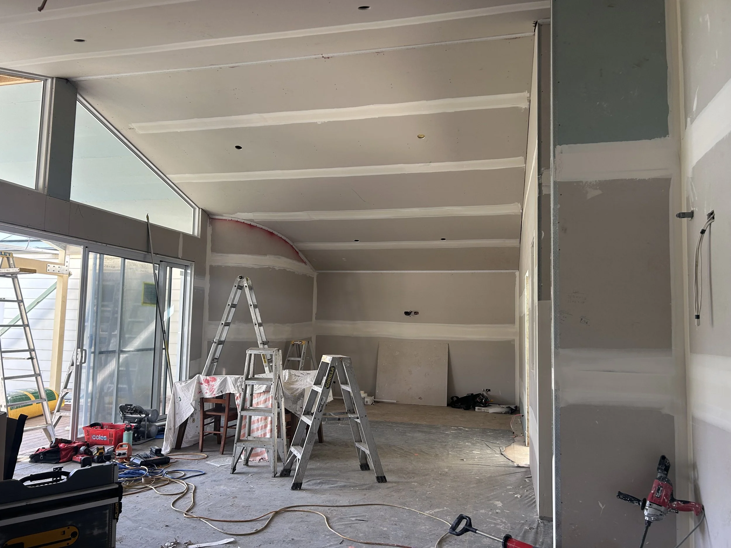Interior of a house under construction with multiple ladders, tools, and construction materials scattered around, drywall partially installed on walls and ceiling.