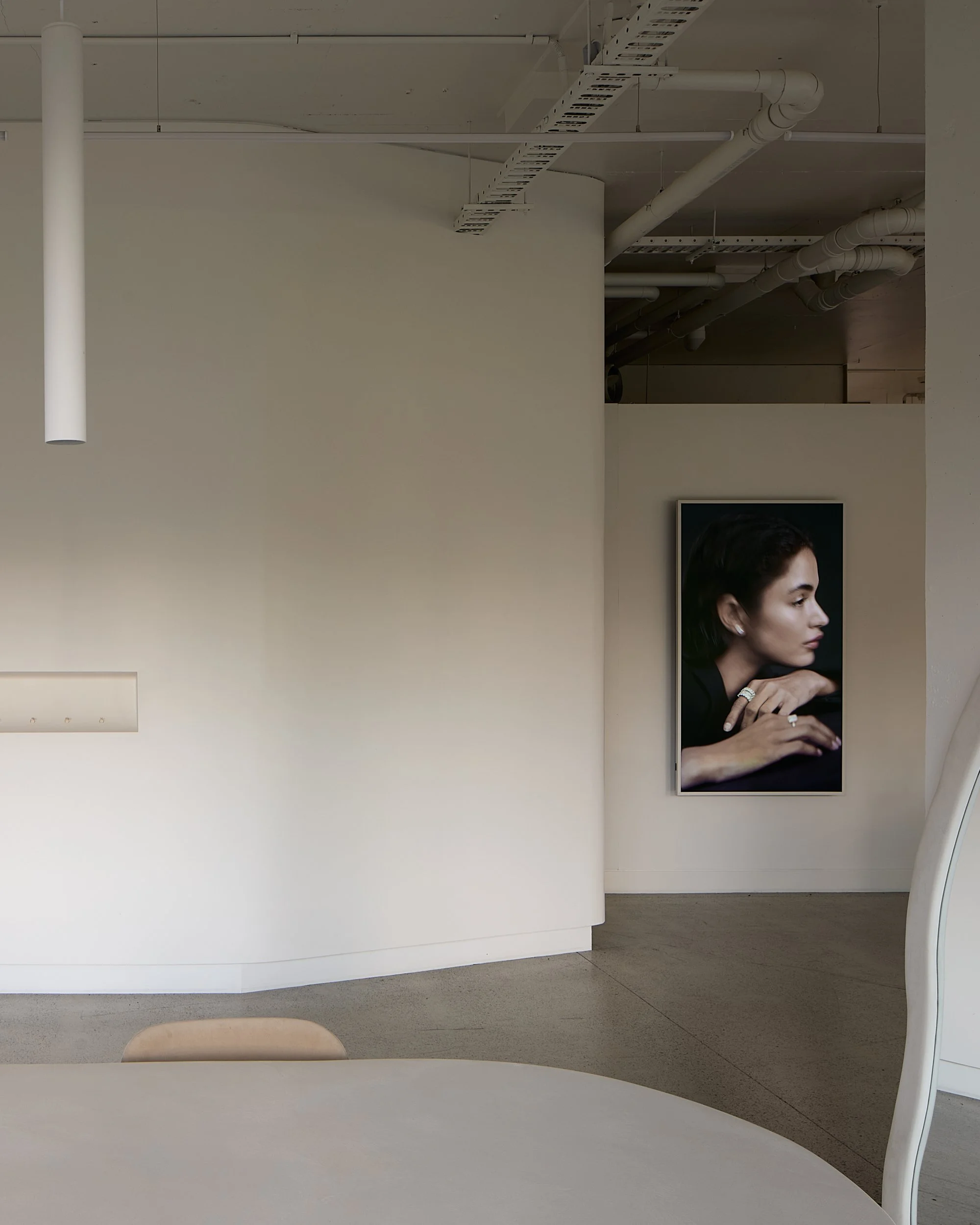 Minimalist interior space with a large framed portrait of a woman with dark hair and makeup, resting her chin on her hand with jewelry, hanging on a white wall.
